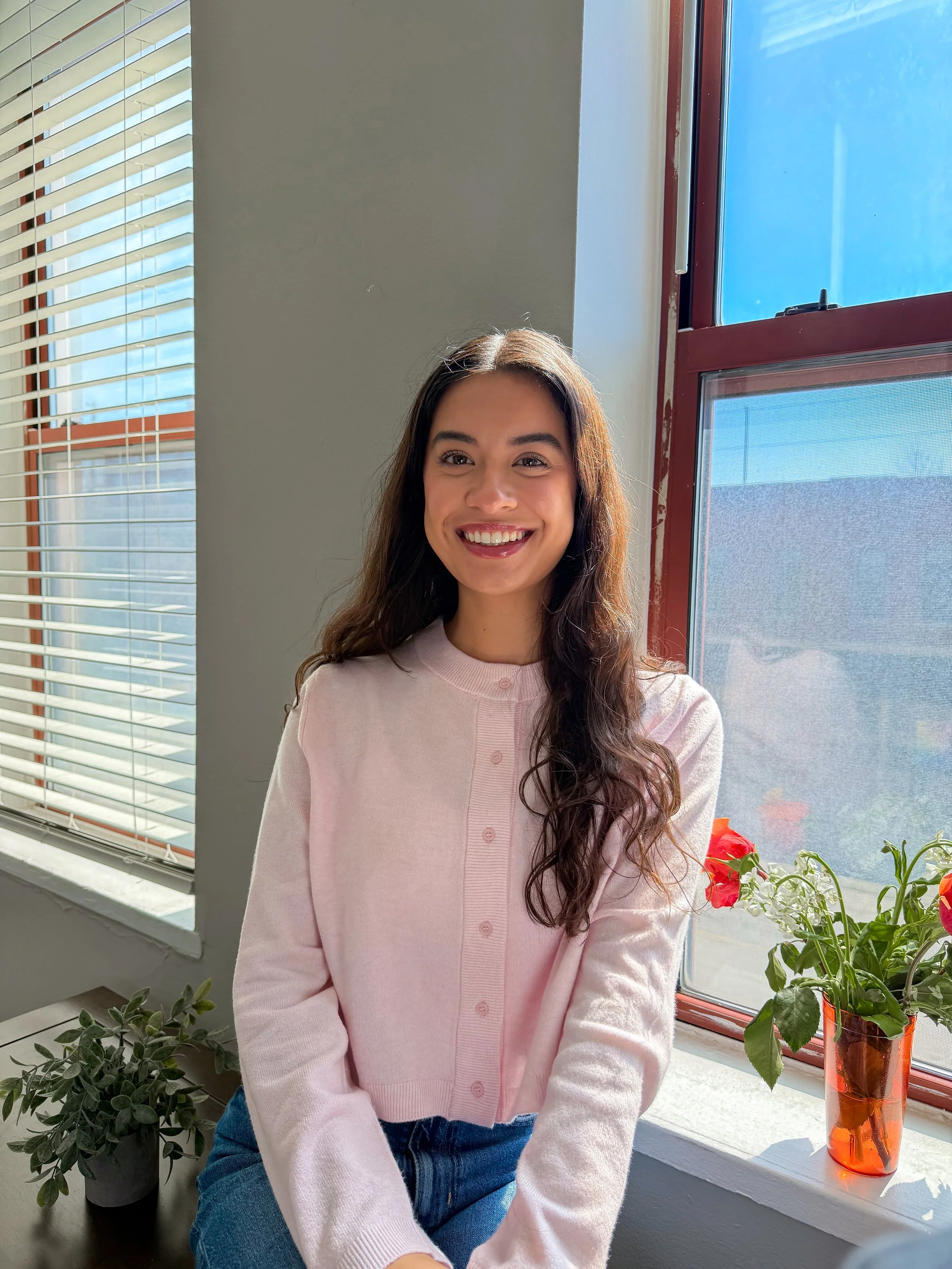 A young woman with long brown hair and a pink cardigan smiling while sitting near a window with blinds, with green plants and red flowers on the windowsill under bright sunlight.