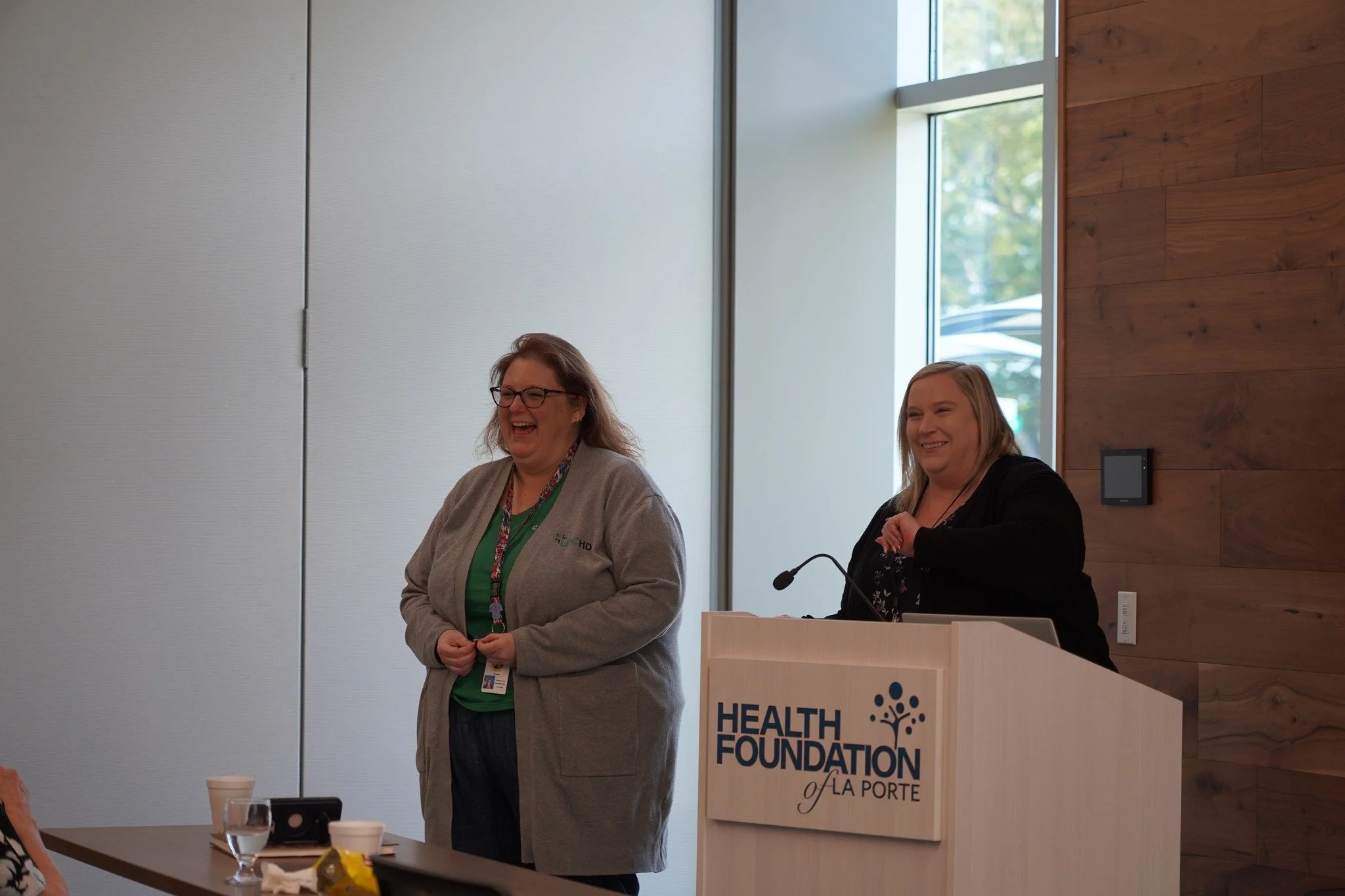 Two women standing and smiling at a podium that reads 'Health Foundation of La Porte,' in a conference room with wood and white walls and large window.