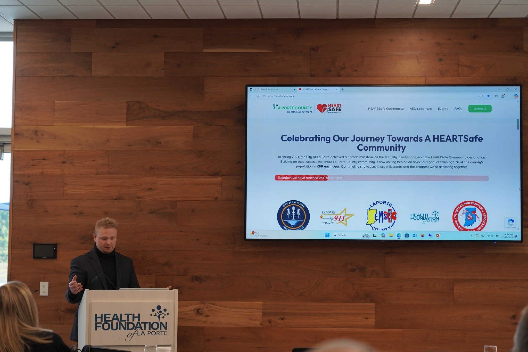 Man giving a presentation at a podium with a sign that says 'HEALTH FOUNDATION of LA PORTE.' Behind him, a large screen displays a webpage from La Porte County Health Department about their journey towards a HEARTSafe community, featuring logos of various local organizations.