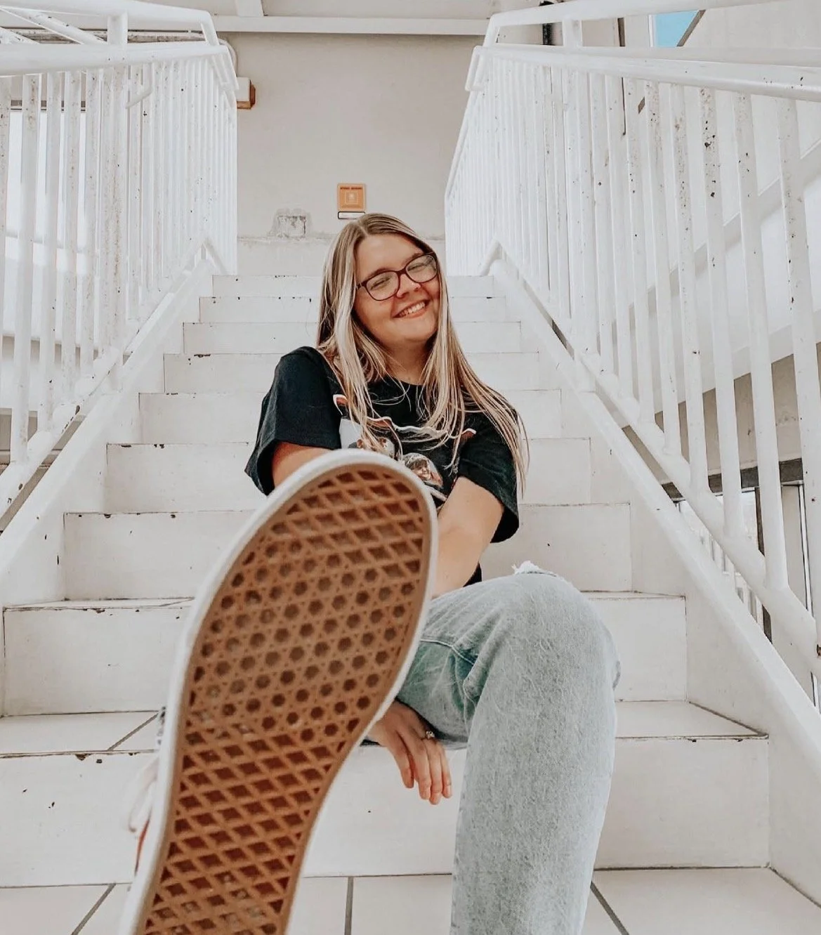 A young woman with glasses and long blonde hair smiling while seated on white stairs, with a shoe in the foreground and white railings on either side.