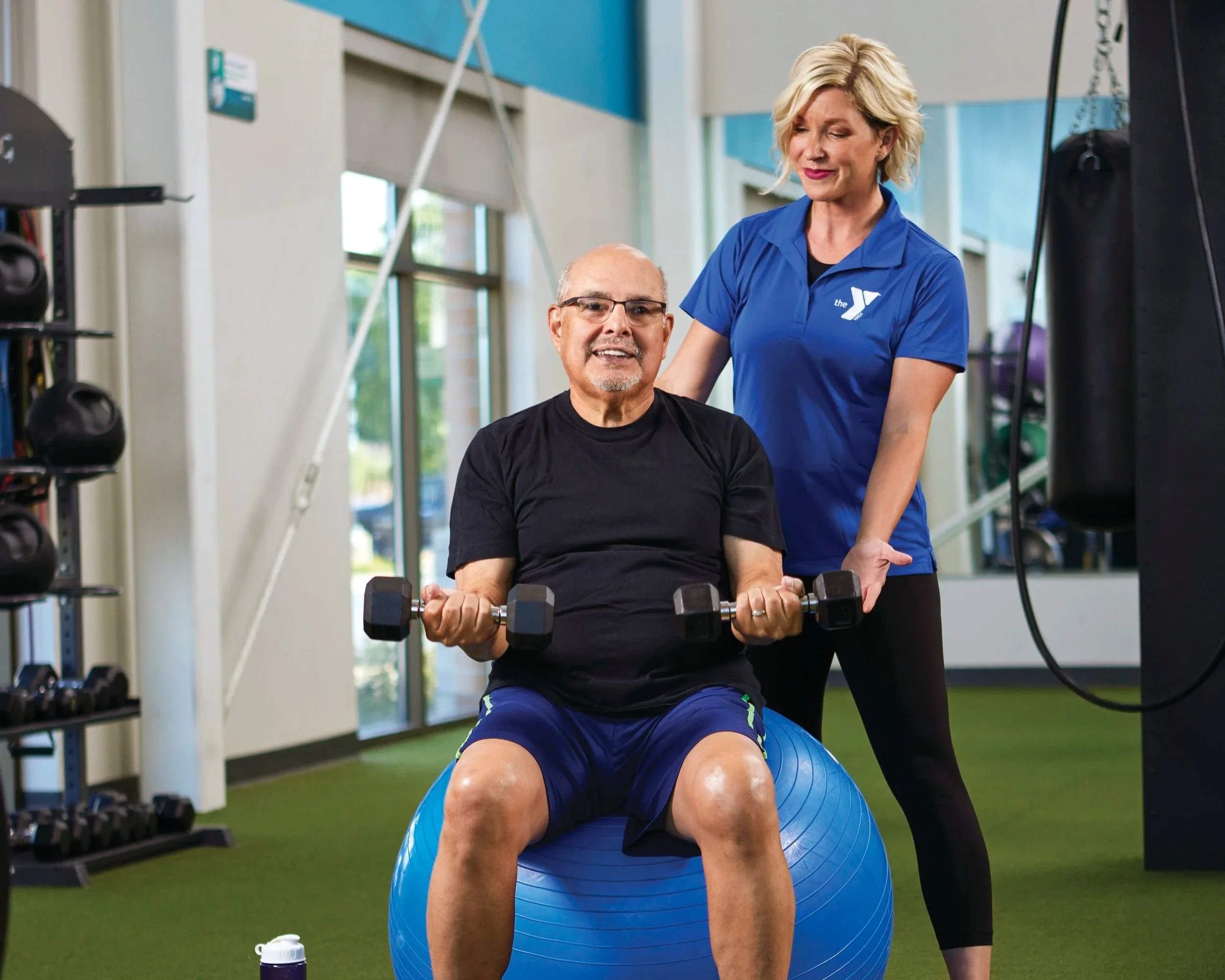 A senior man exercising with dumbbells on a blue exercise ball at a fitness center, assisted by a female trainer wearing a blue YMCA polo shirt.