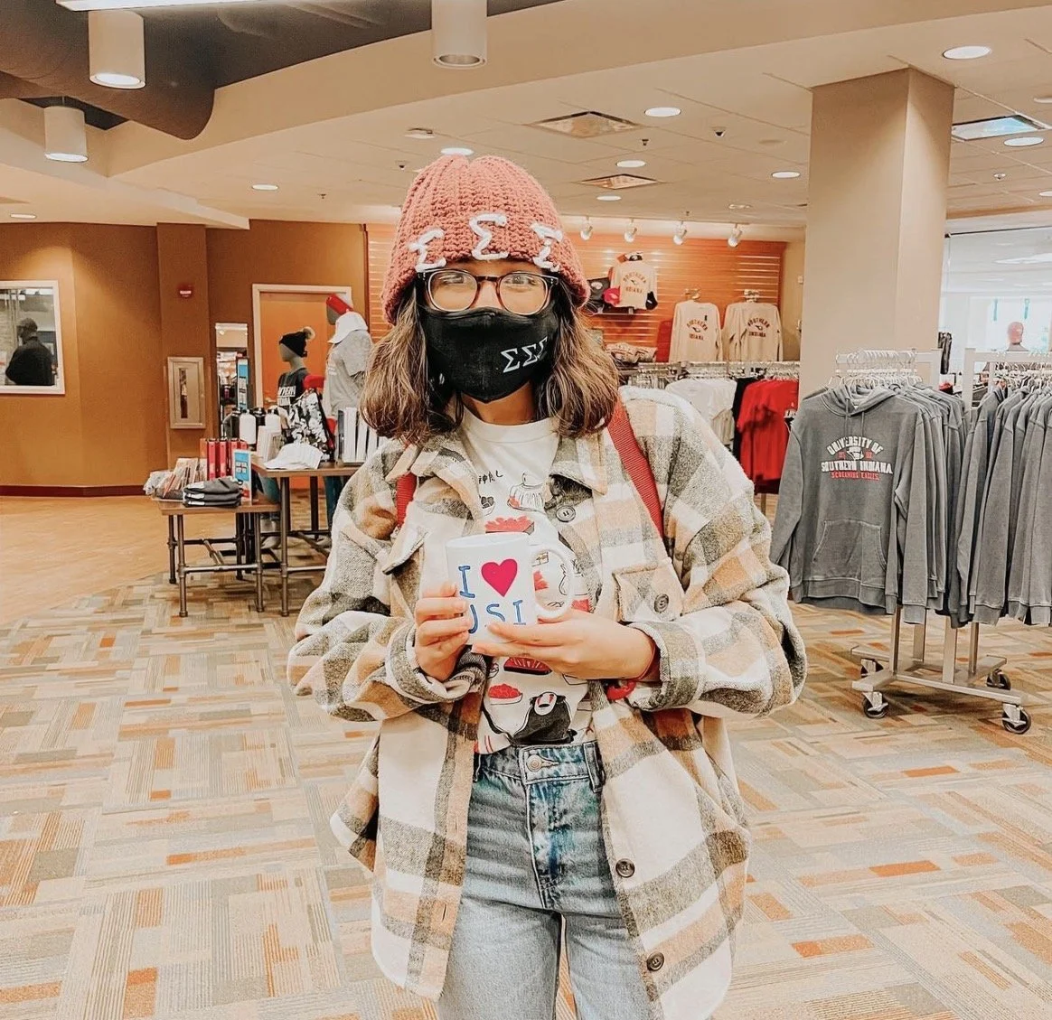 A young woman with glasses, a black face mask, and a pink knit hat with a Sigma fraternity symbol, standing inside a store with sports apparel and merchandise. She is holding a cup with writing and hearts on it, wearing a plaid jacket over a graphic 