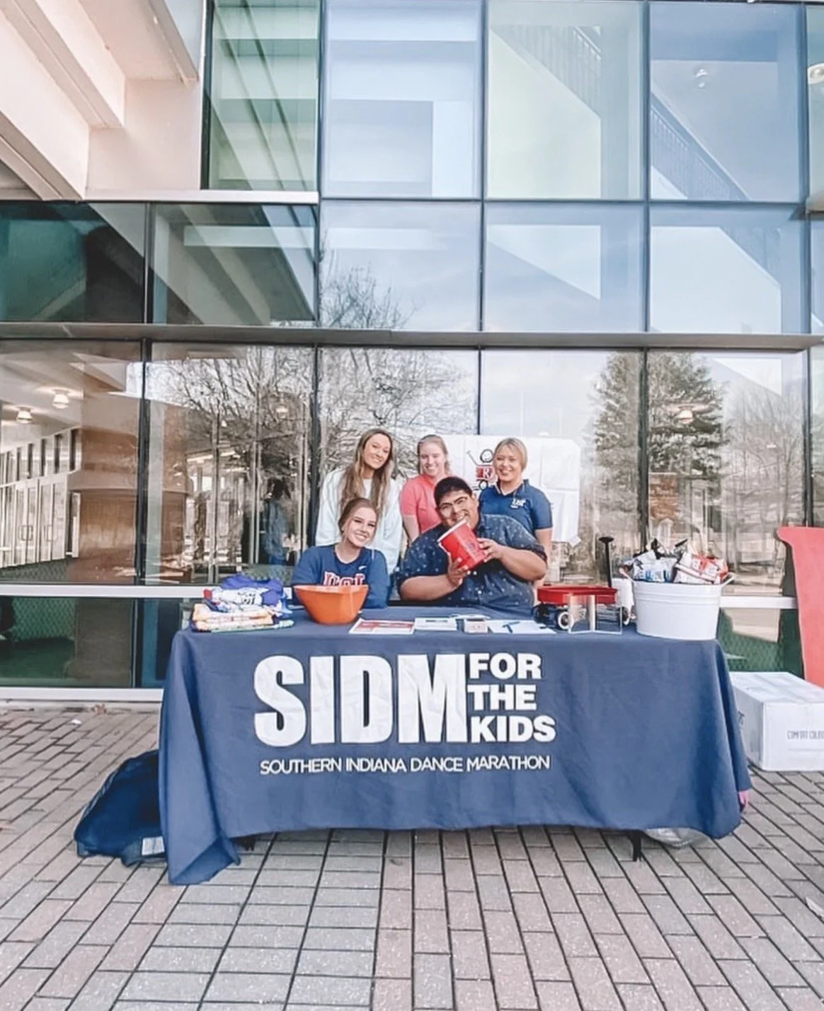 Group of five young people at a table outside a building with glass windows, promoting the Southern Indiana Dance Marathon for the kids, with various items and donation containers on the table.