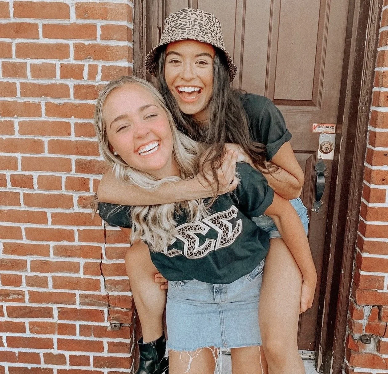 Two young women smiling and hugging outside a brick building, one woman carrying the other on her back near a wooden door.