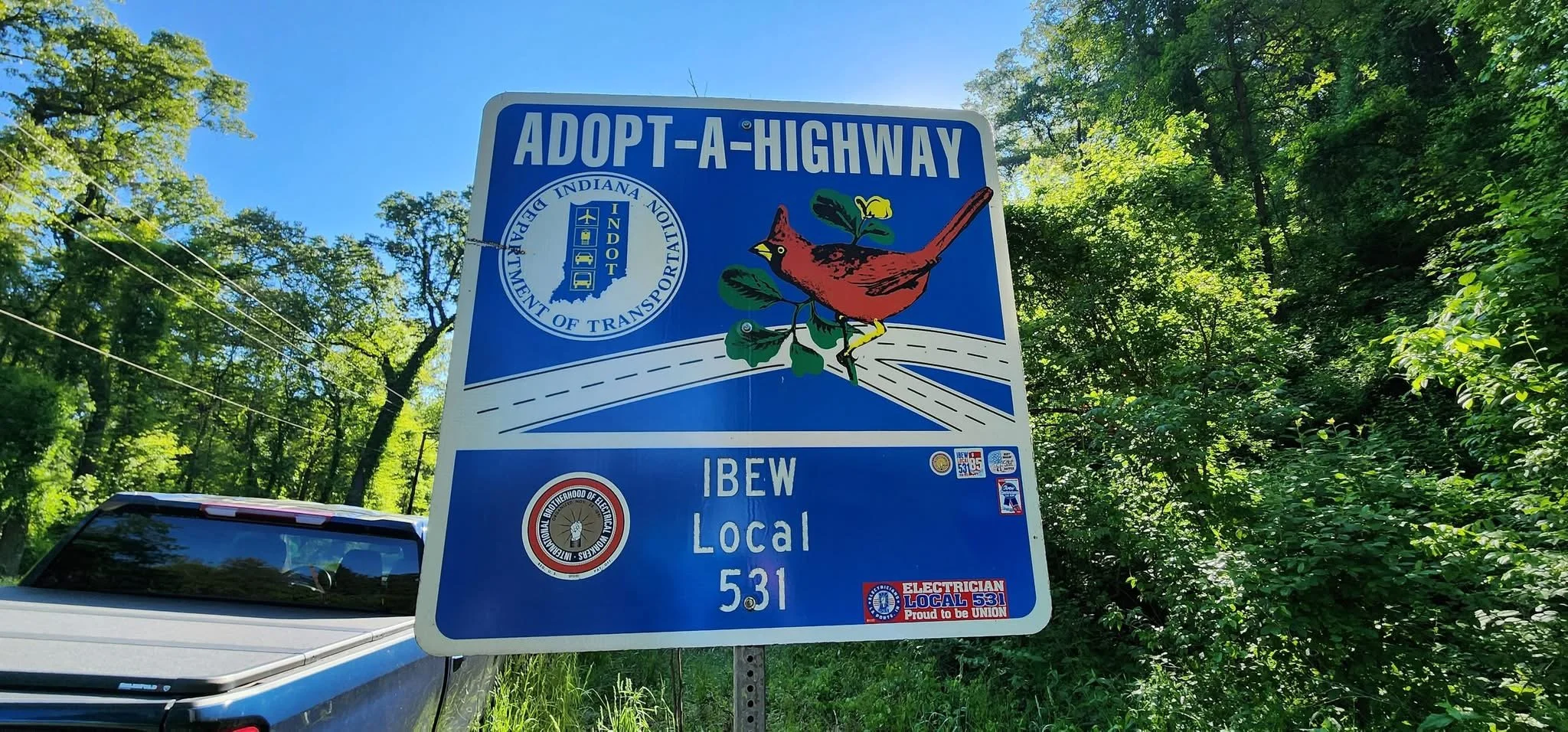 Blue highway sign with the words "IBEW Local 531 Adopt-A-Highway" and a colorful bird illustration, surrounded by green trees and a parked vehicle in the background.