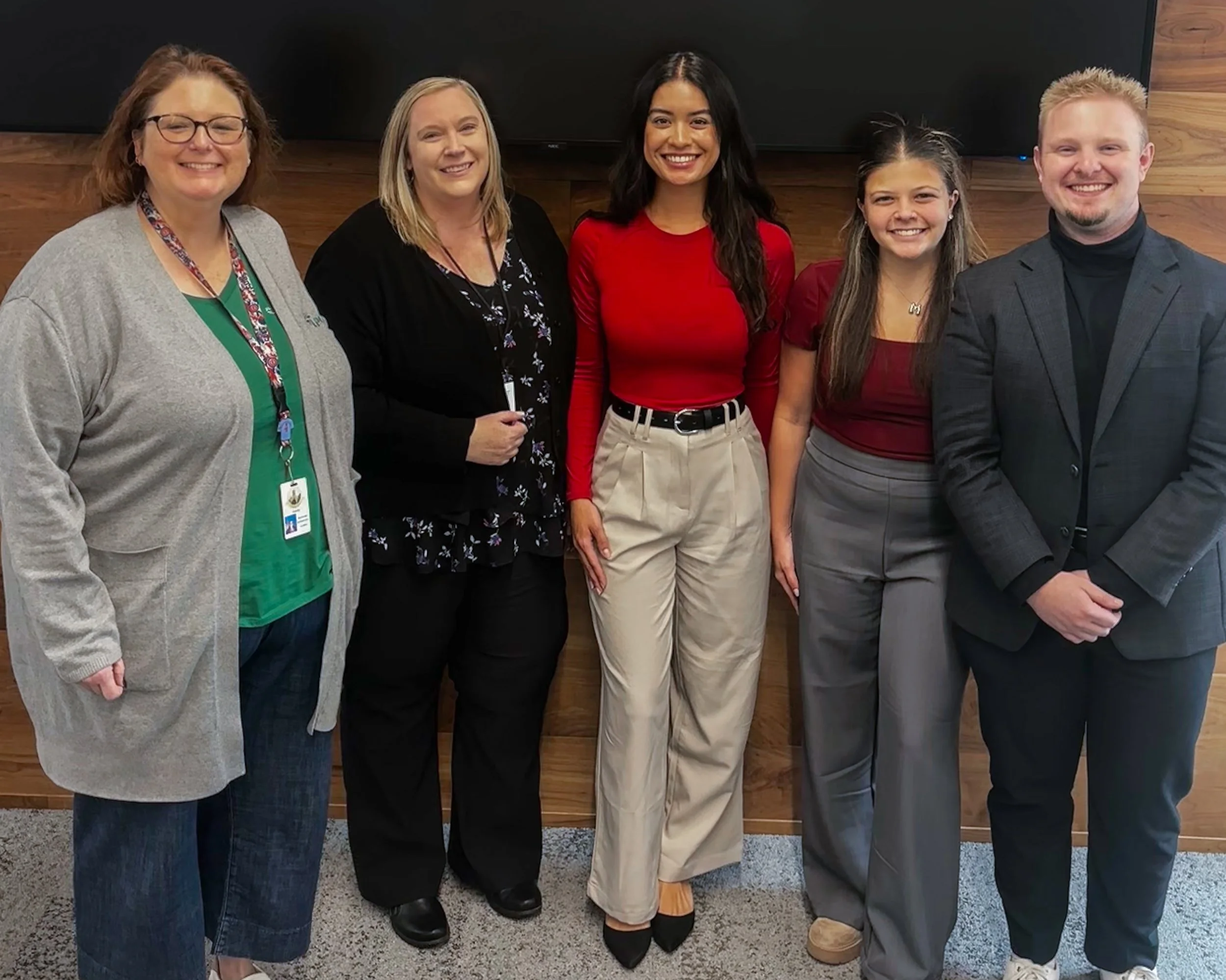 Group of five people standing together in an office, smiling for a photo. They are dressed in casual and business attire, with a wooden wall and a black screen behind them.