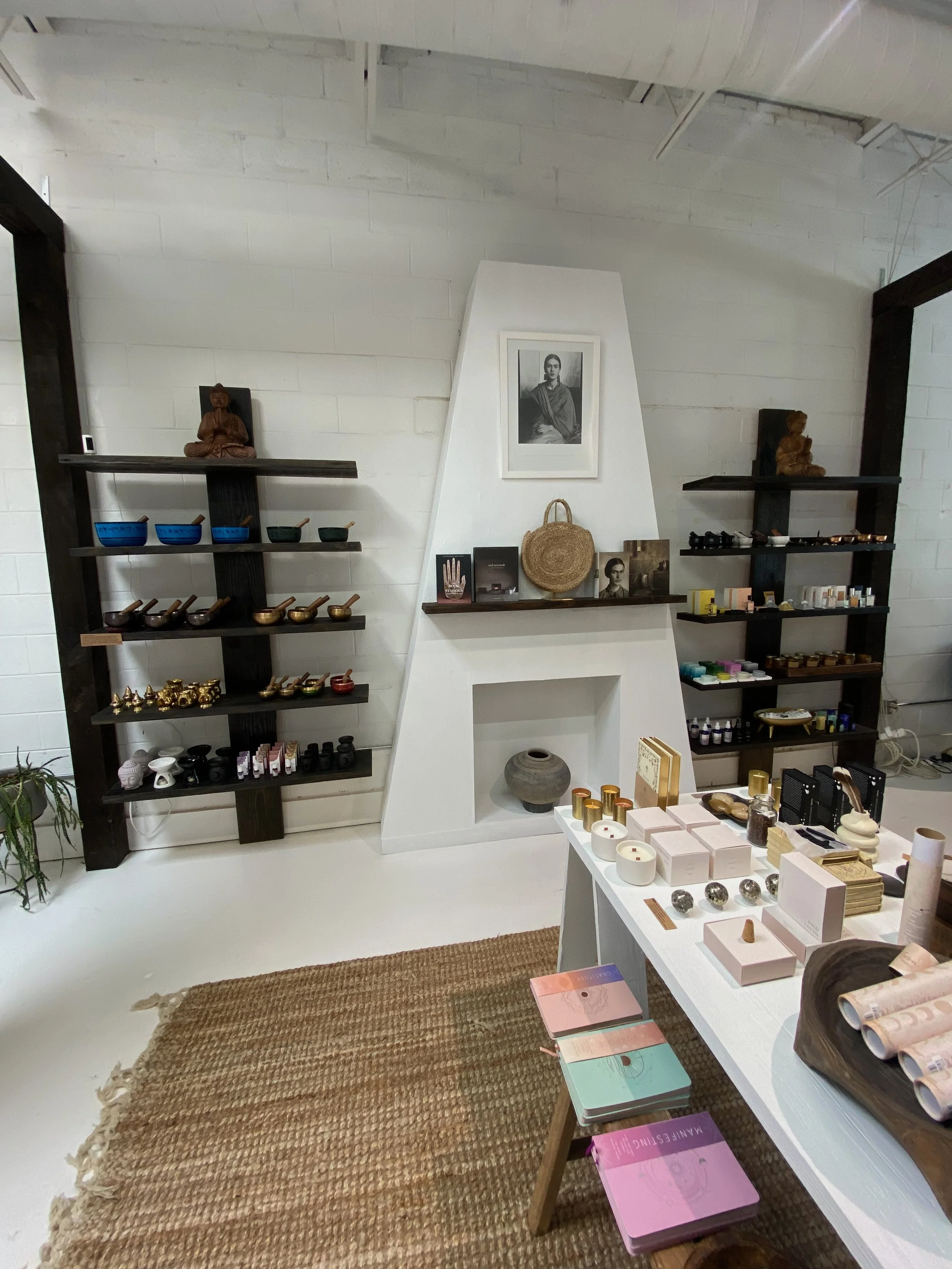 Interior of a retail shop with shelves displaying various small items such as candles, soaps, and decorative objects. There is a white table in the foreground with pastel-colored notebooks and other products. A woven rug is on the floor.