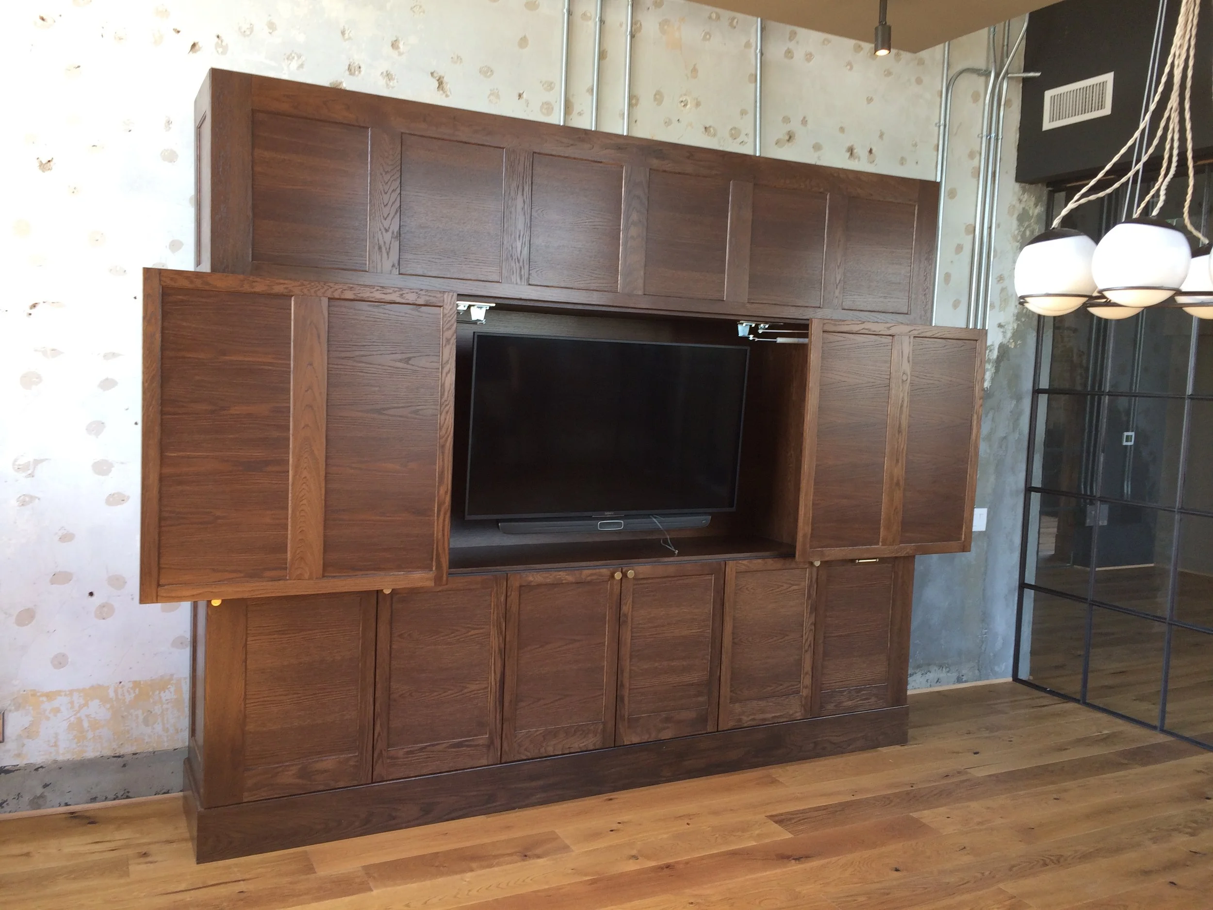 A large wooden entertainment center with multiple cabinets and open space, housing a flat-screen TV, situated against a wall with peeling white wallpaper. To the right, there is a black metal shelving unit and a modern lamp with round white shades hanging from the ceiling.