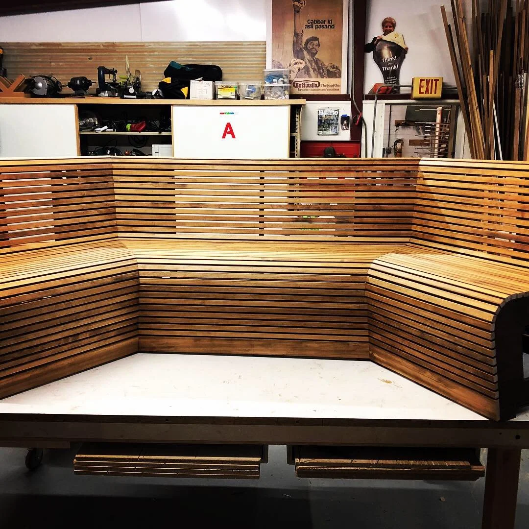 Unfinished wooden L-shaped bench with slatted design in a workshop setting, with tools, posters, and storage in the background.