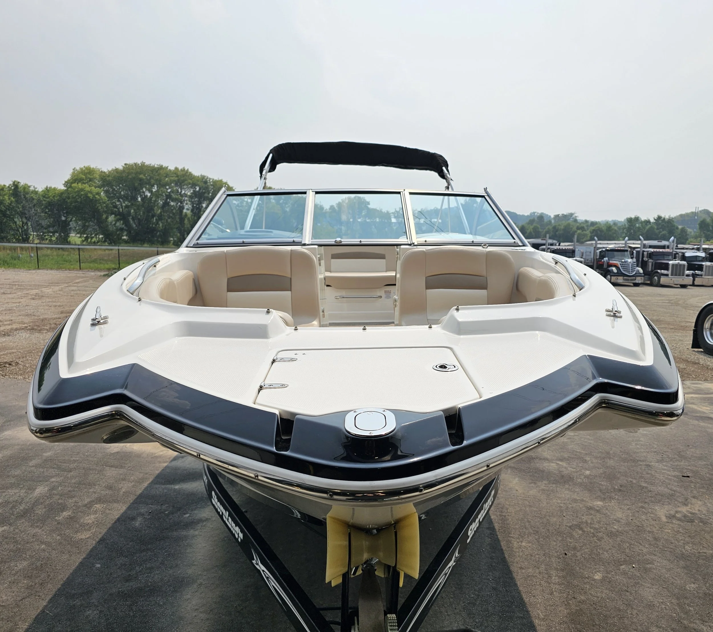 Front view of a white and black speedboat with beige interior seats, parked outdoors with a line of trucks in the background.