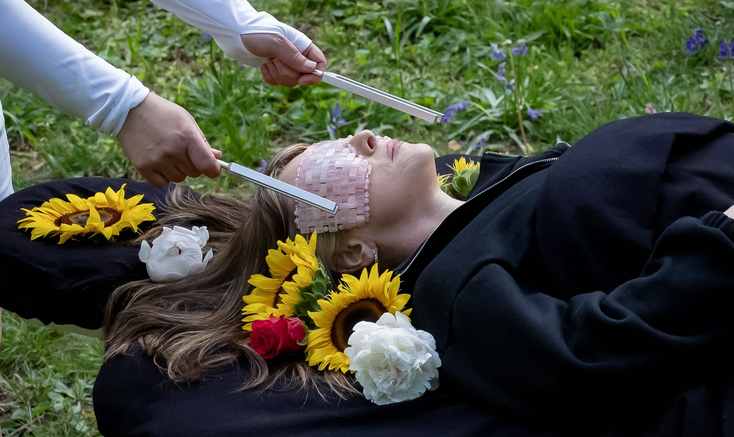 A woman with a bandaged forehead lying on the ground with her eyes closed, surrounded by flowers. Someone is holding two metal objects near her head, possibly as part of a medical or ritual procedure. The setting appears to be outdoors on grass.