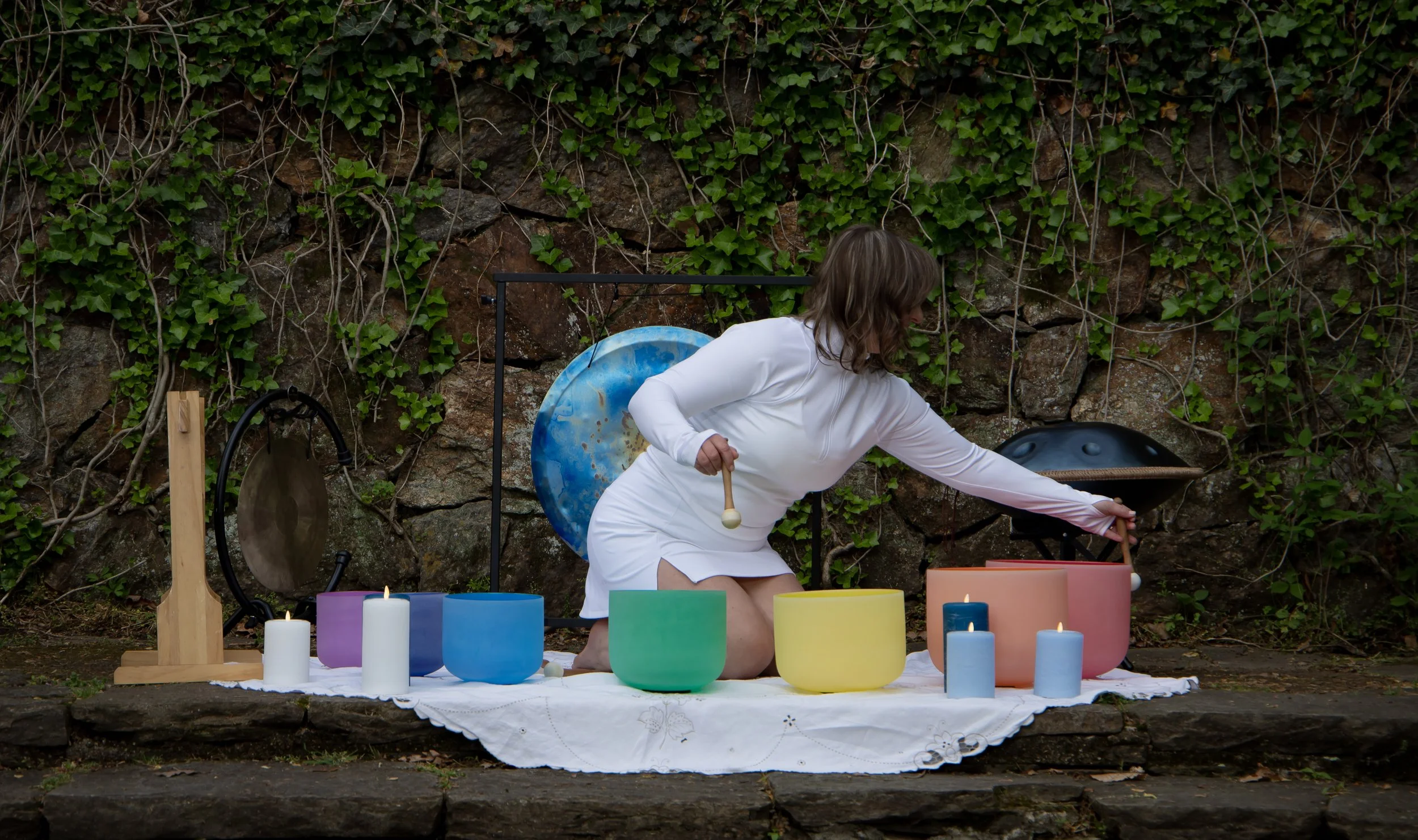 A woman dressed in a white outfit performing a musical or spiritual ritual outdoors, surrounded by colorful candles and musical instruments such as a gong and a hang drum, against a rocky wall covered with green ivy.