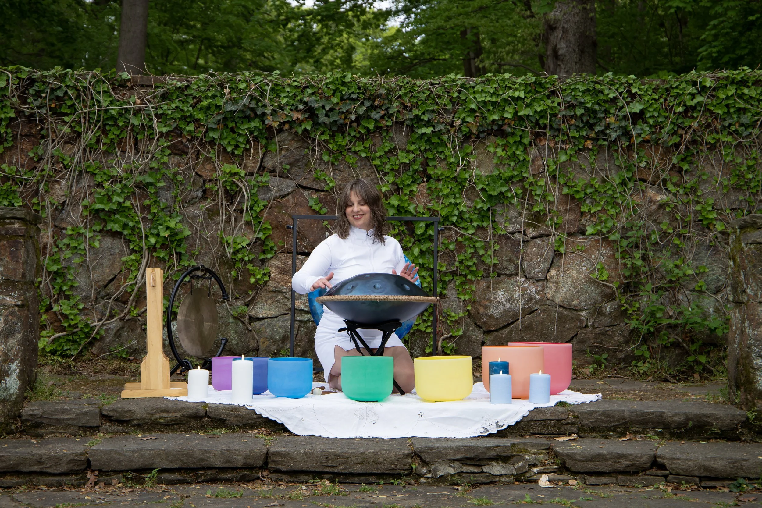 A woman smiling and sitting outdoors in front of a stone wall covered with green ivy, playing a handpan drum. In front of her, there are colorful candles, bowls, and a gong.