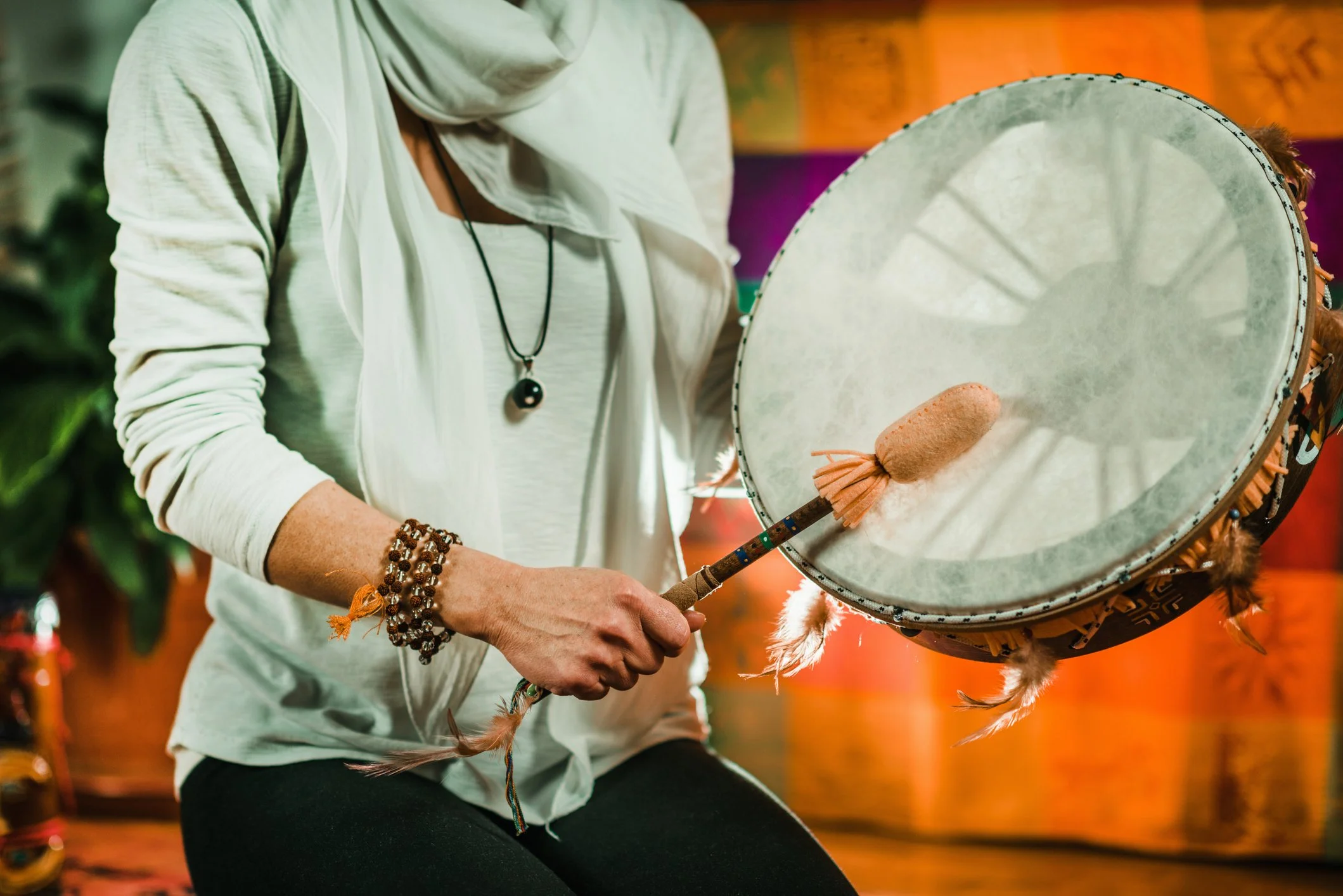 Person playing a drum with a beaded bracelet on their wrist, wearing a white shirt with a scarf. The drum has feathers attached to it.