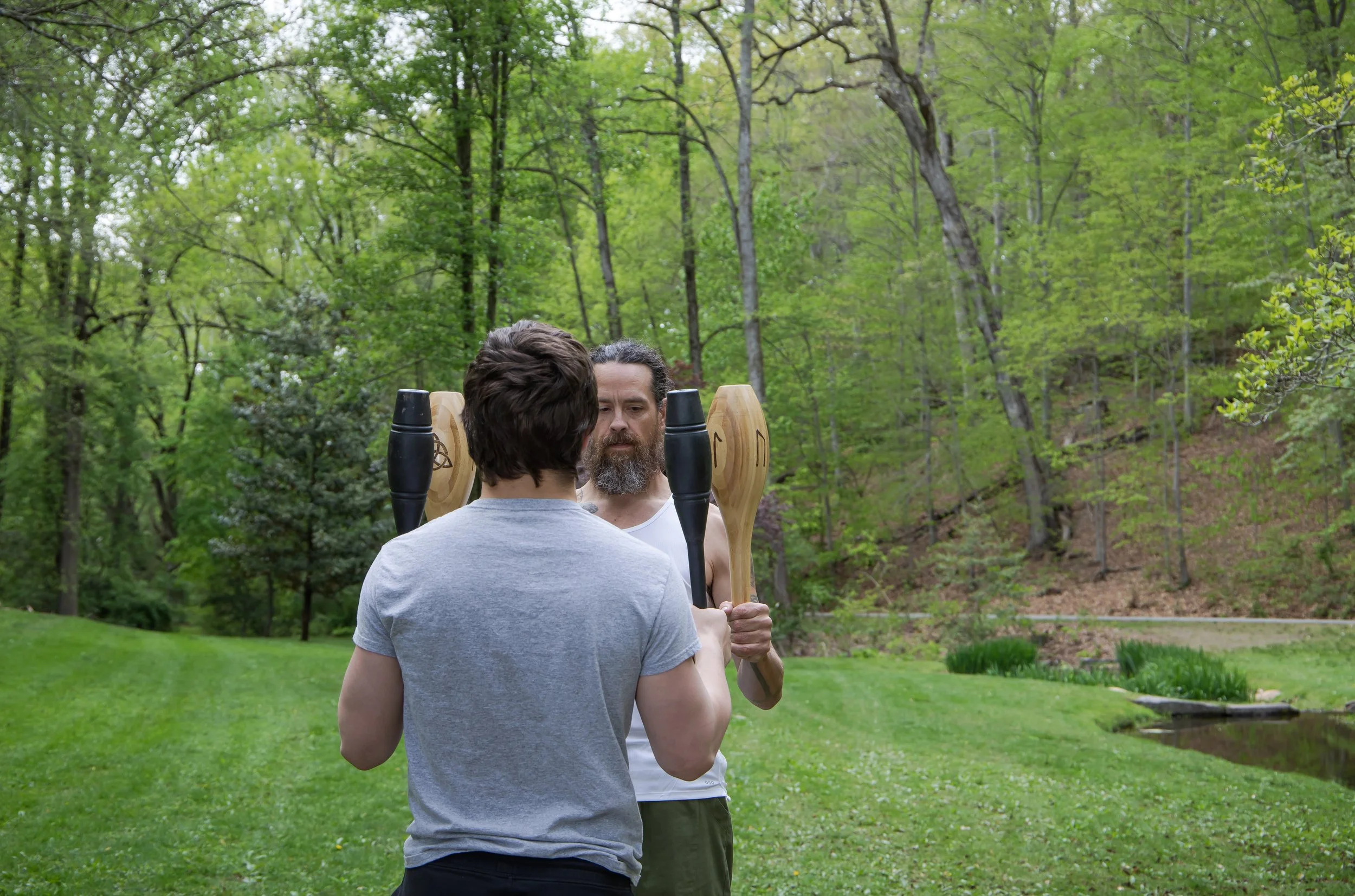 Two men practicing martial arts with wooden training weapons outdoors in a lush green park, surrounded by tall trees and grass.