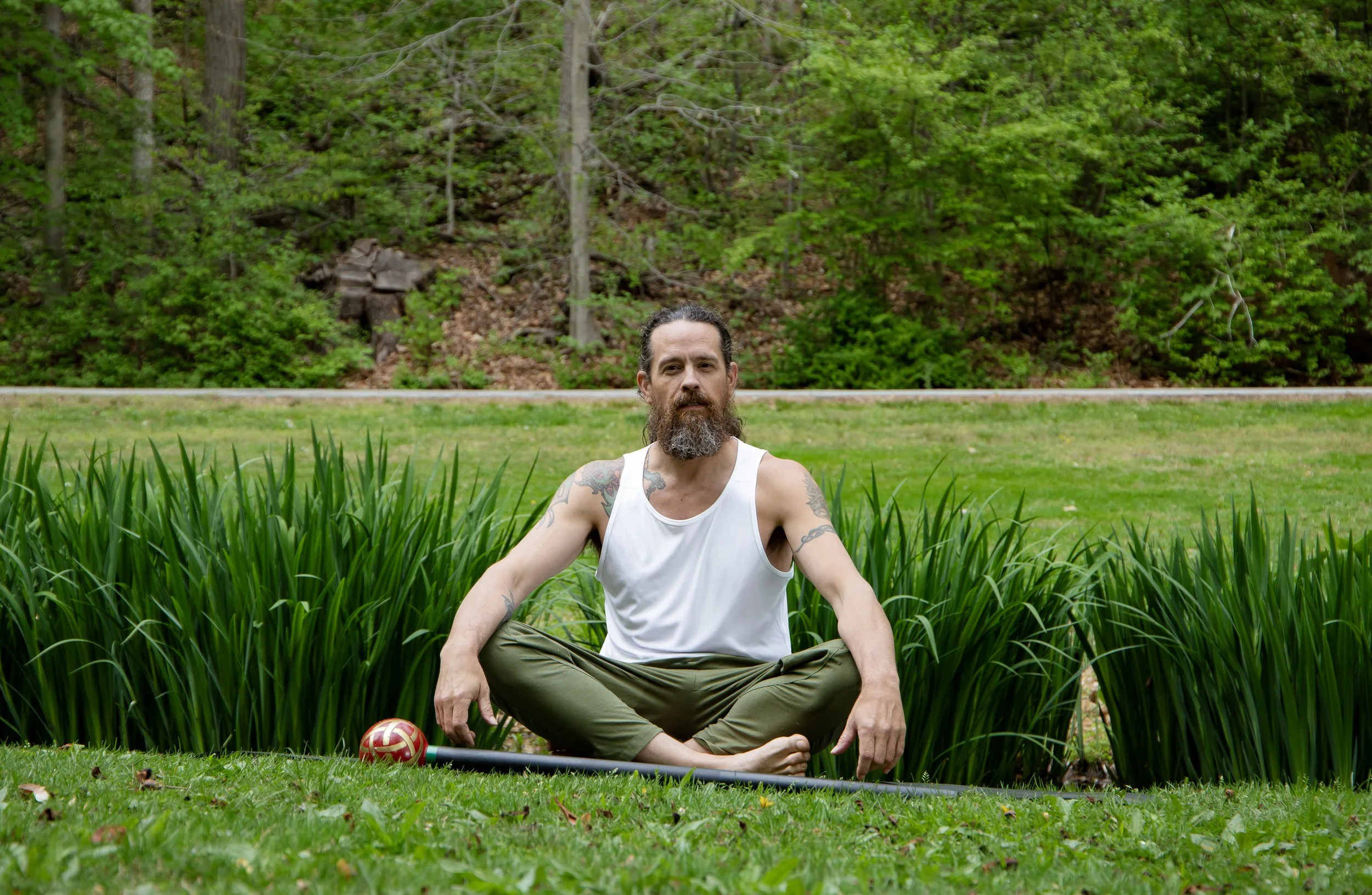 A bearded man in a white tank top and olive pants sitting cross-legged on the grass in a park or garden. He is meditating with eyes closed, surrounded by green plants and trees in the background.