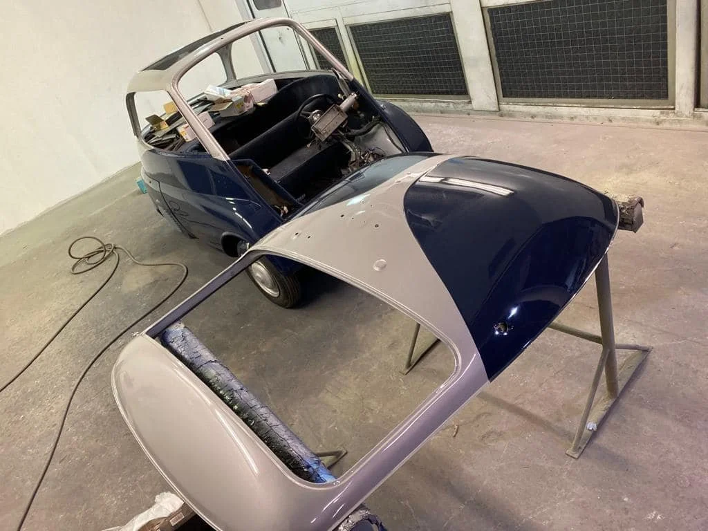 A vintage car in the process of restoration with a blue body and a gray hood, placed on a stand with two support bars in an indoor garage or workshop.
