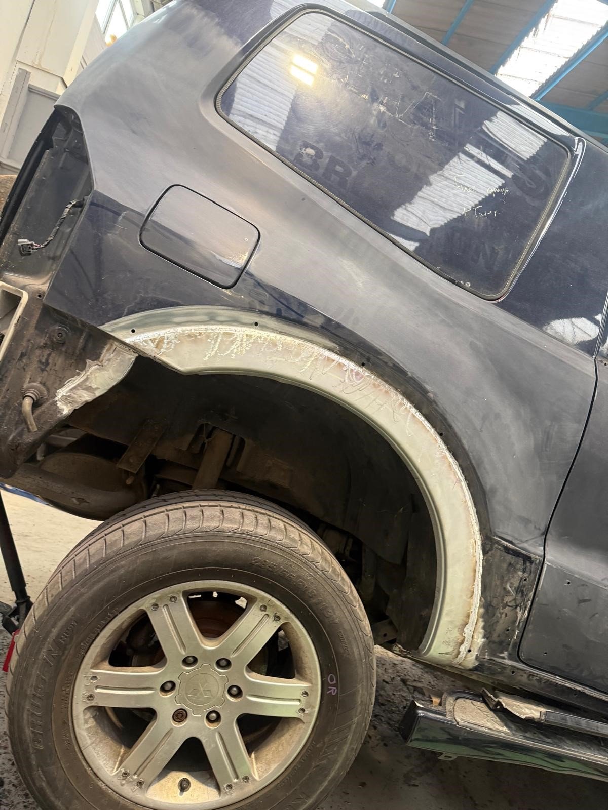 Front left side of a car with a missing front wheel, showing wheel well and bodywork restoration in progress, with a workshop background.