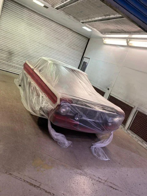 A car covered with transparent plastic in a garage or workshop, with a partially visible red stripe on its side and covered wheels.