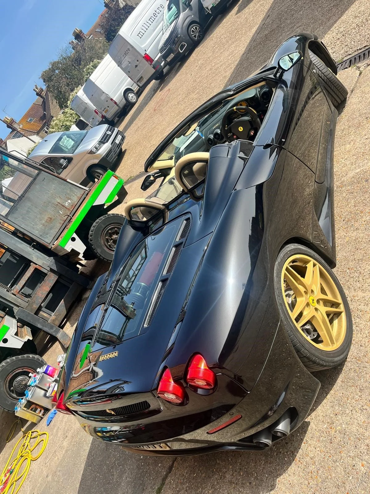 A black Ferrari sports car with gold wheels parked on a street, with several vans and trucks nearby, some with green accents, and a cityscape in the background.