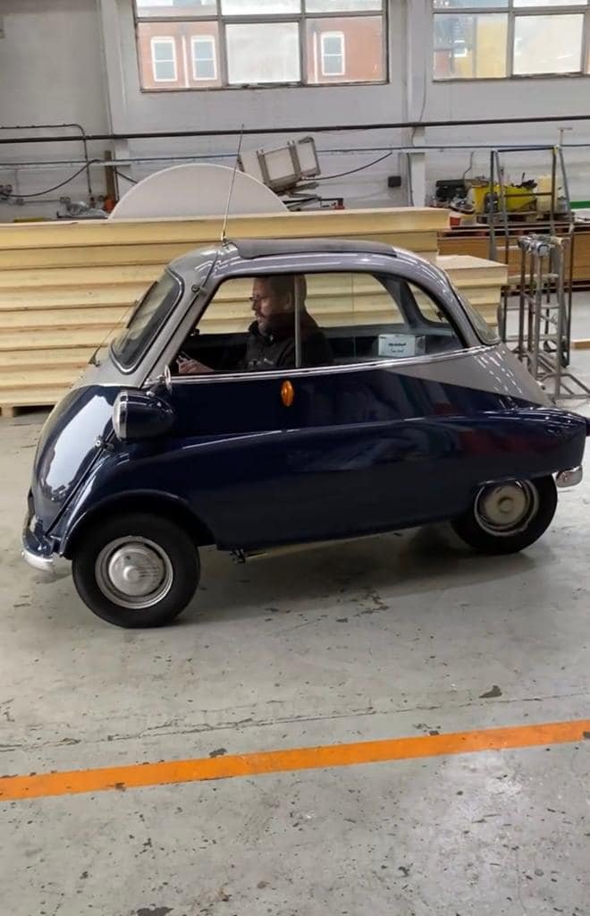 A man inside a small, vintage-style, black and gray toy car or miniature vehicle in an industrial workshop or warehouse.