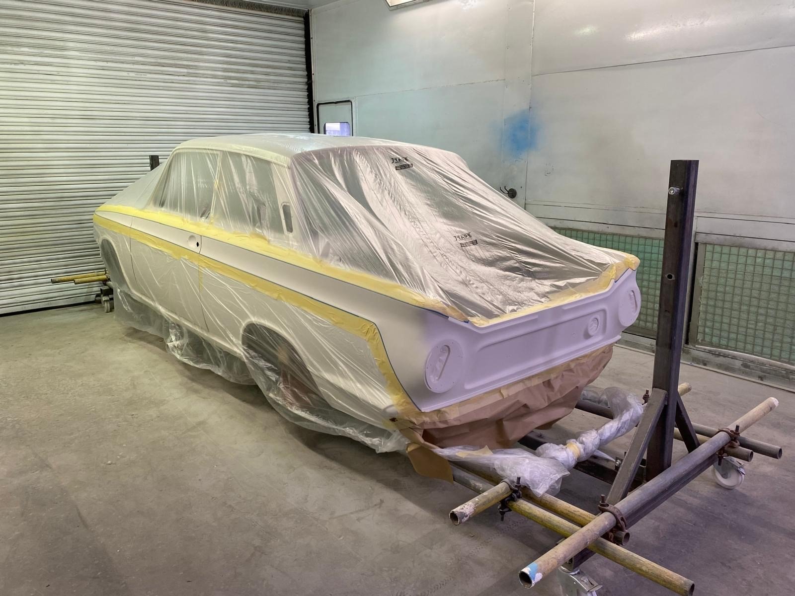 A car body in a paint shop, covered with protective plastic sheeting, mounted on a scaffolding frame.