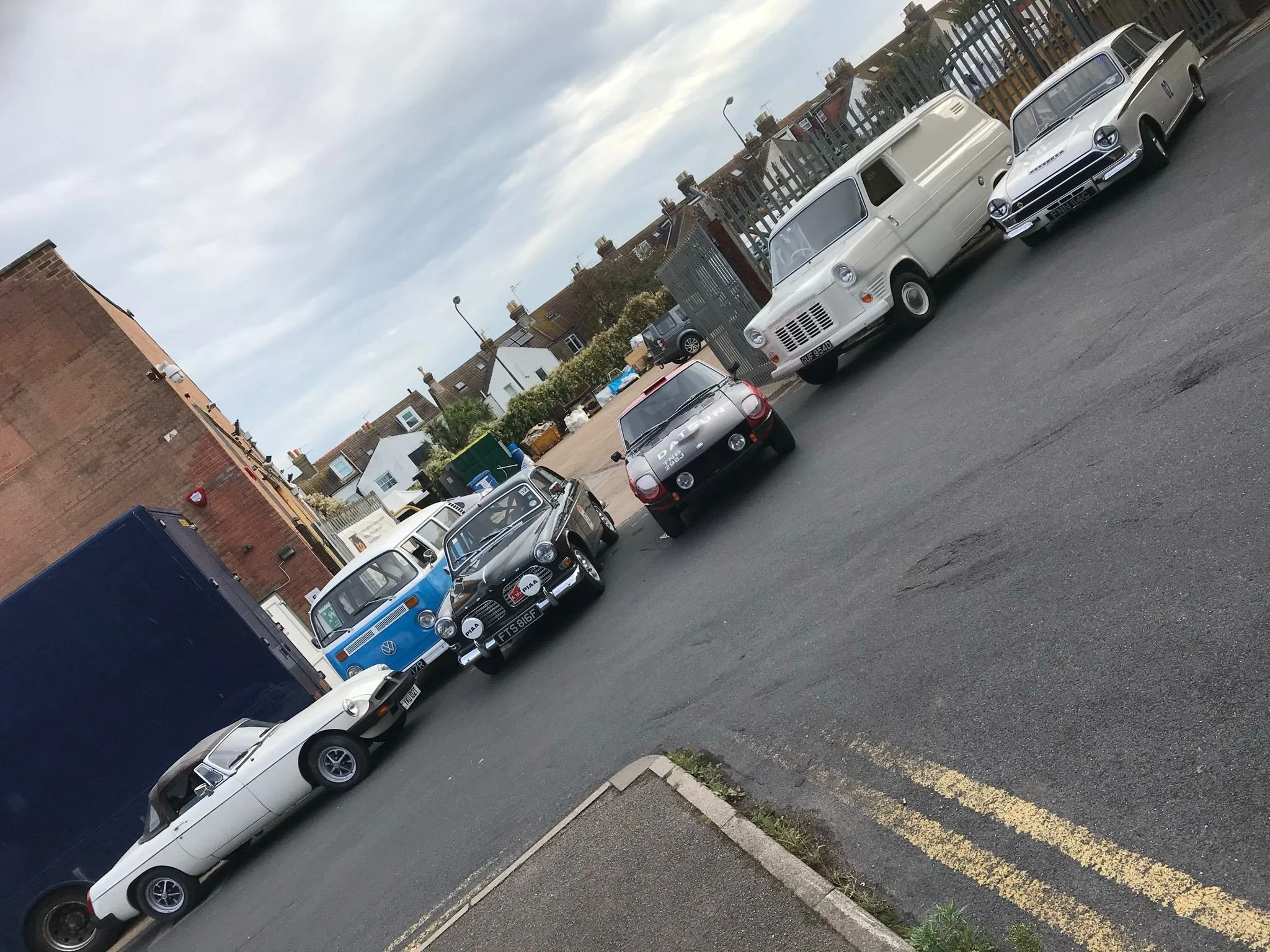 A lineup of vintage and classic cars parked along a residential street with houses and a fence in the background under cloudy skies.