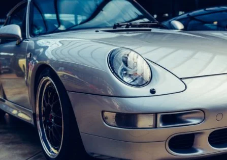 Close-up view of a silver sports car, showing the front headlight, part of the hood, and wheel with a black rim, parked outdoors.