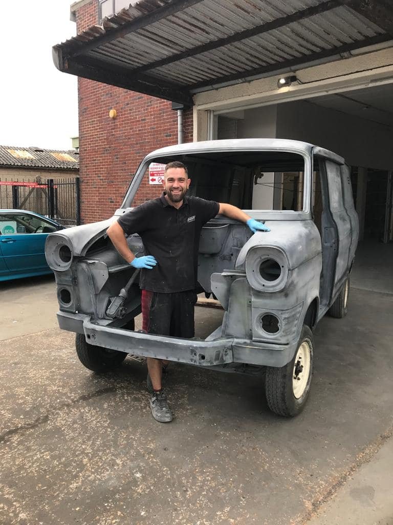 A man wearing a black shirt, shorts, and blue gloves standing next to a vintage car body shell in a workshop or garage.