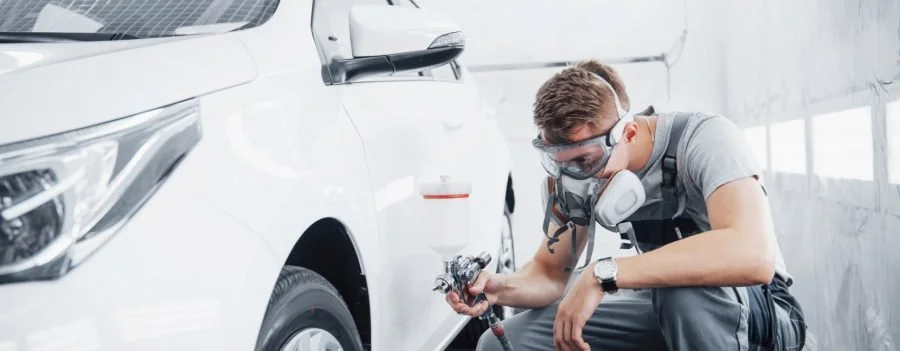 A man wearing safety goggles, a respirator mask, and gray clothing is spray painting a white car in a workshop.