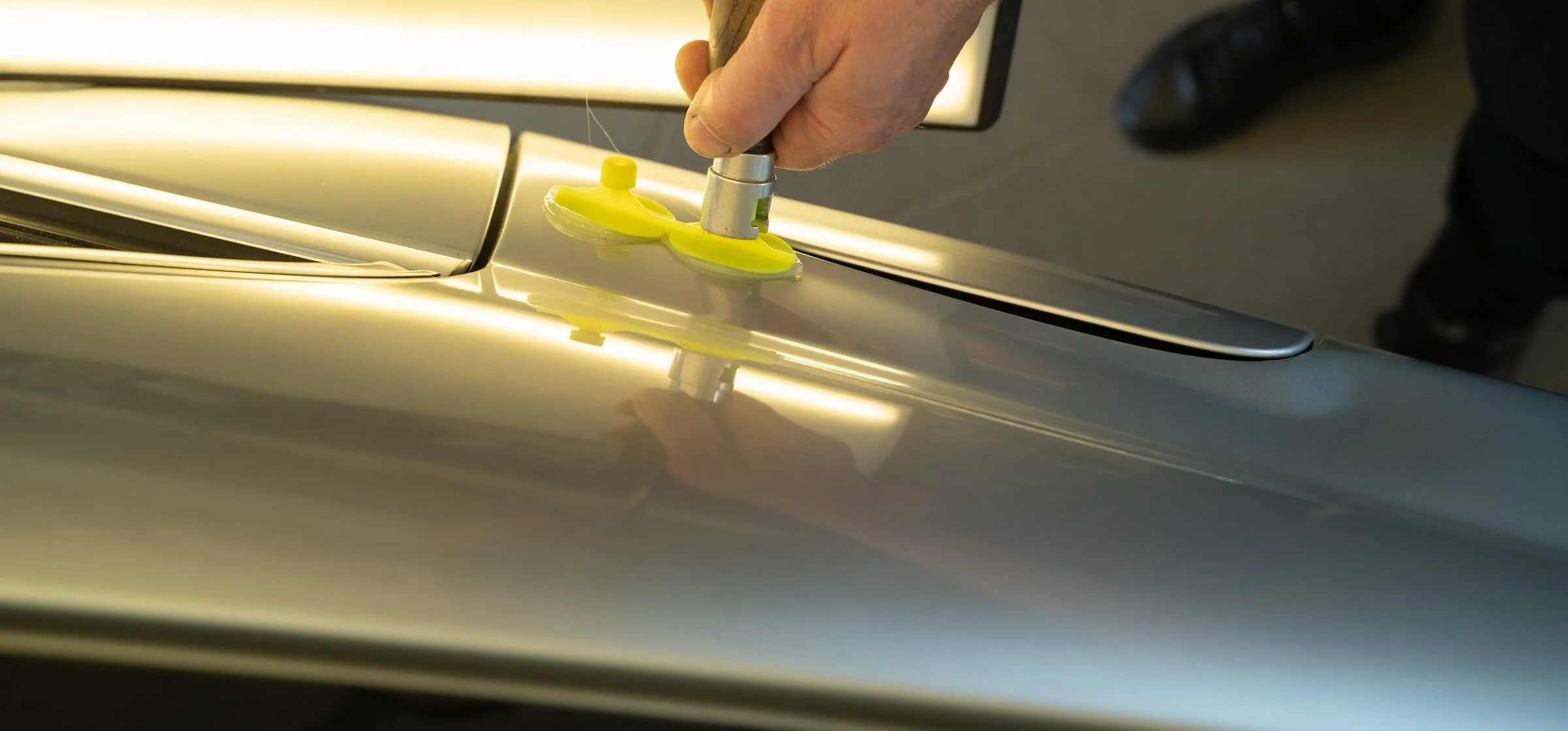 A person using a yellow suction cup tool to repair a shiny silver car's surface.