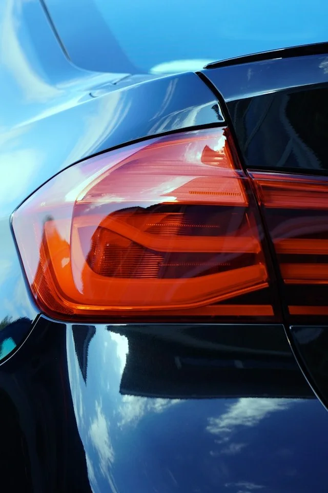 Close-up of the rear left taillight and part of the trunk of a black car, with reflections of the cloudy sky on the shiny surface.
