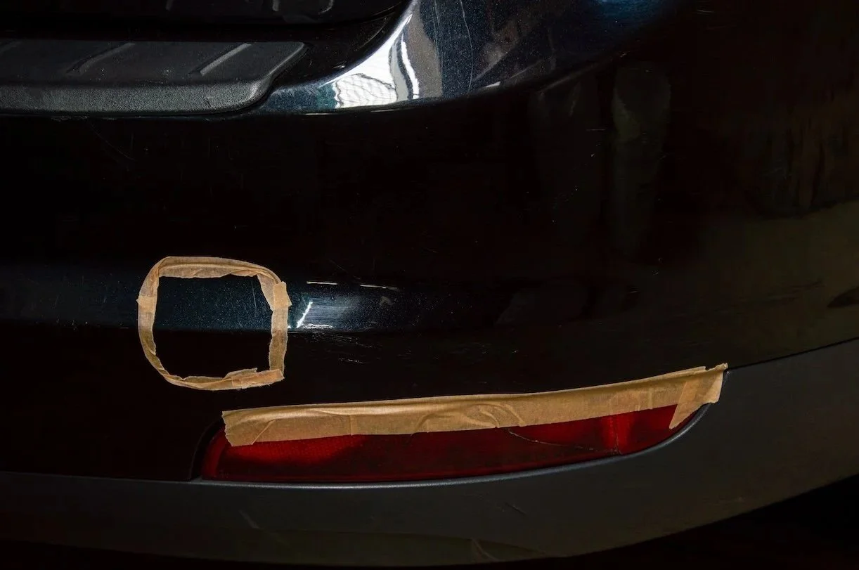 Close-up of a black car's rear bumper with masking tape around a large circular and a long horizontal section, likely for repair or painting work.