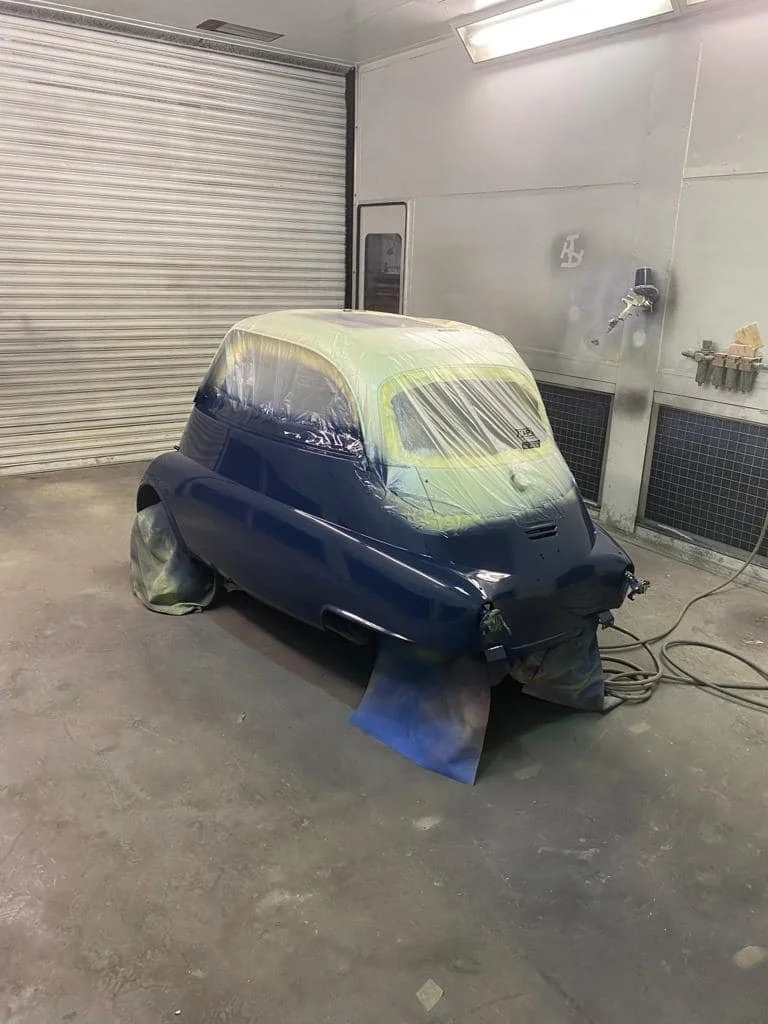 Small vintage car being painted, with the body covered in protective plastic and masking tape, inside a workshop.