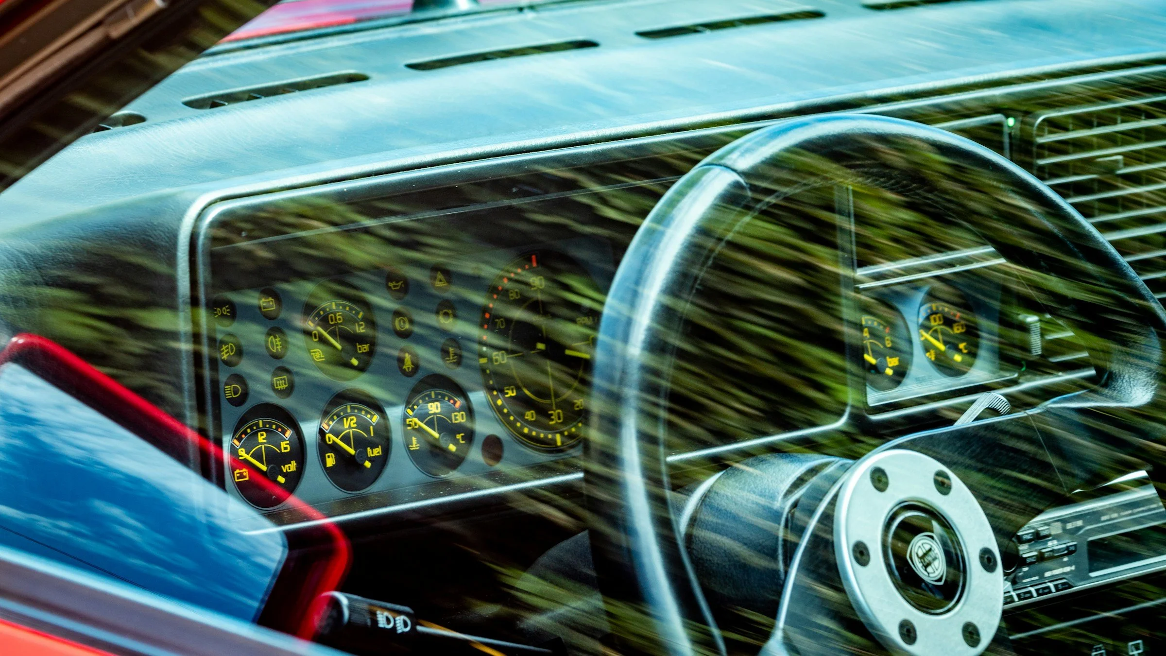 Close-up view of a vintage car dashboard with analog gauges, steering wheel, and reflection of trees outside.