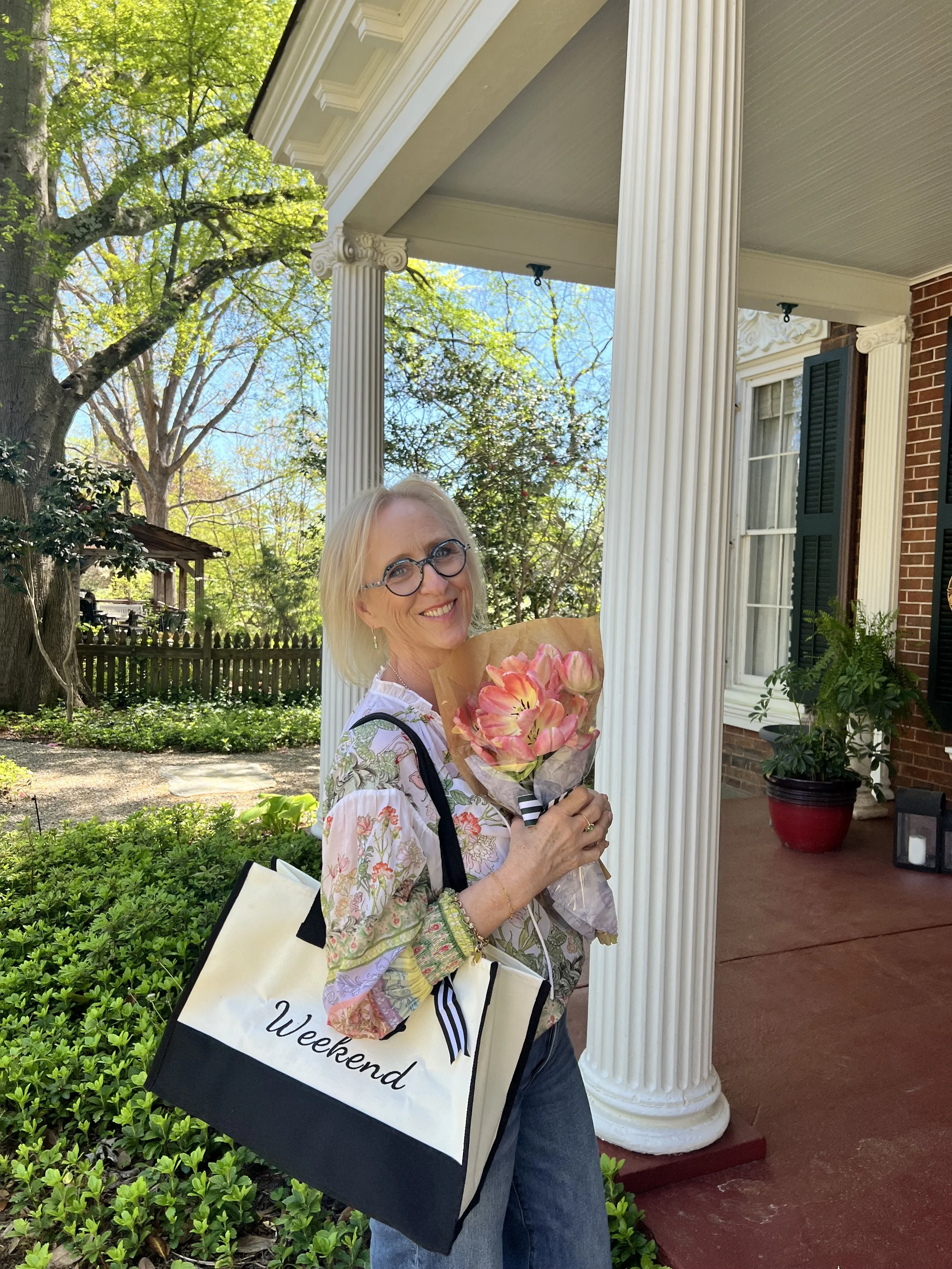 A woman with glasses, blonde hair, and a floral blouse holding a bouquet of pink flowers, standing outside a house with a front porch, white columns, and a lush garden with trees and greenery.