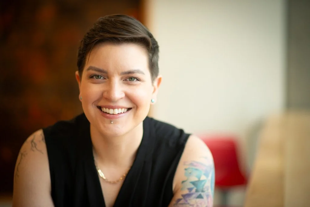 A smiling woman with short dark hair, tattoos on her arms, facial piercings, wearing a black sleeveless top, sitting indoors.