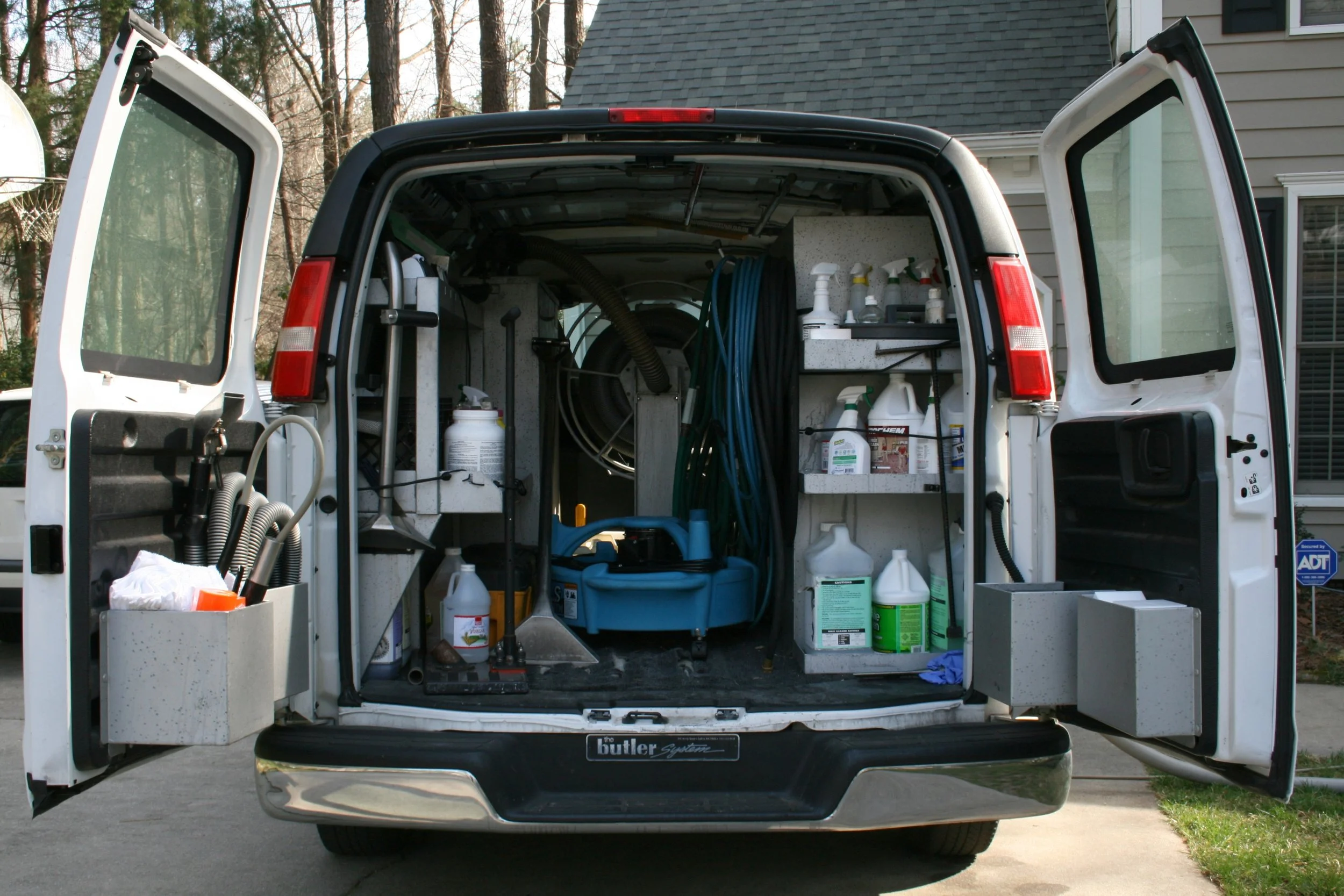 The back of a Mumin Janitorial Services utility van with open doors, showing organized cleaning and maintenance supplies, including spray bottles, hoses, and cleaning chemicals.