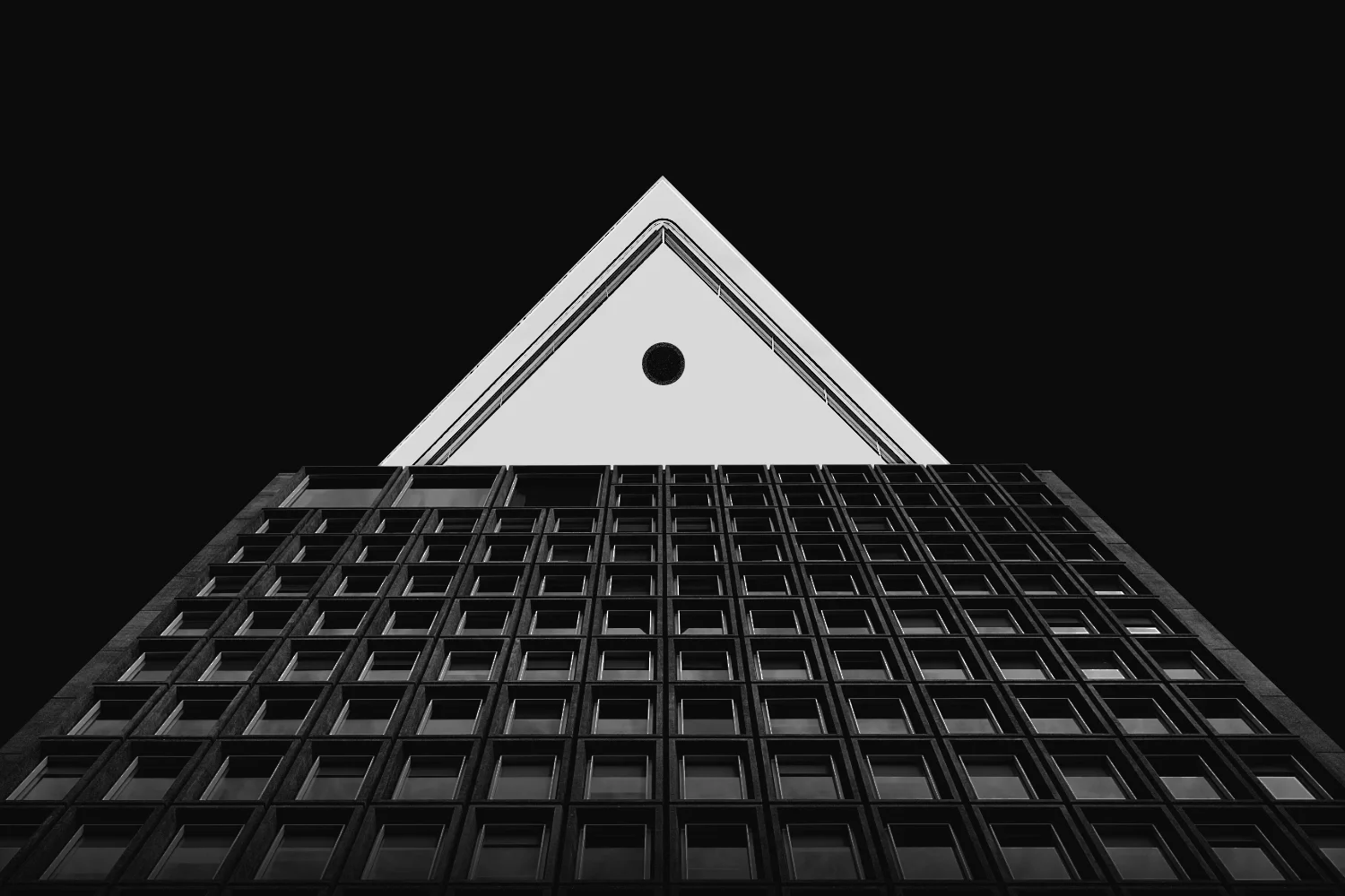 Low angle view of a modern high-rise building with a grid of windows, against a dark sky and a white triangular structure at the top.