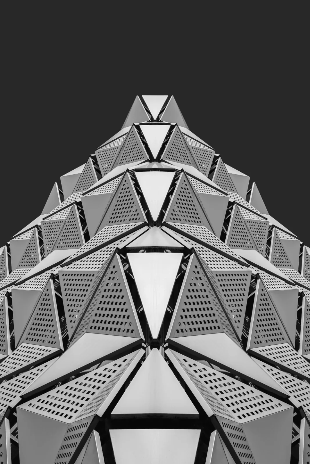 Black and white photo of a modern, geometric building with a pyramid-like structure, featuring multiple triangular and rectangular panels with perforations, viewed from below against a dark sky.