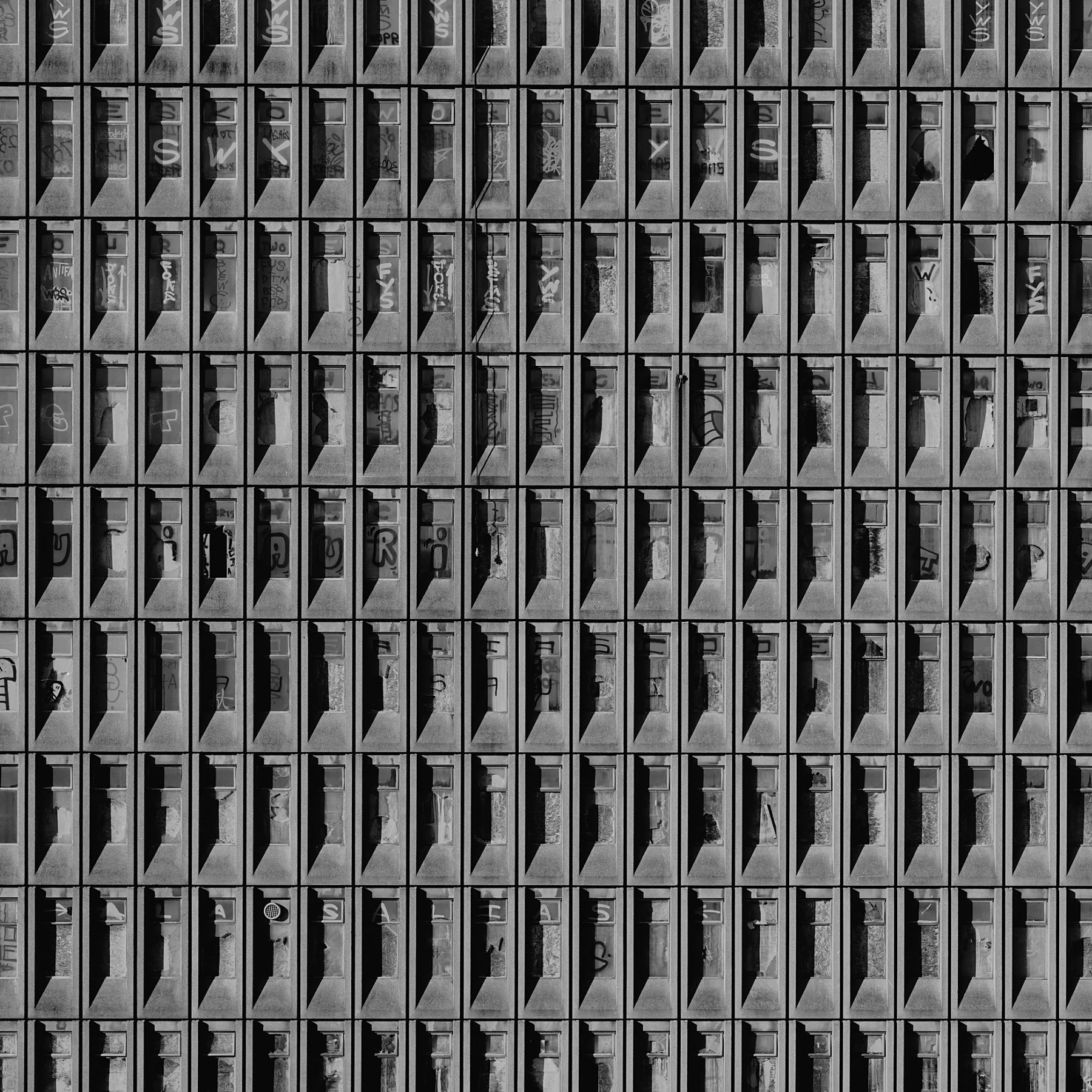 A close-up black and white photograph of a building's grid of concrete balconies with glass railings, some of which have graffiti and objects such as chairs and plants.