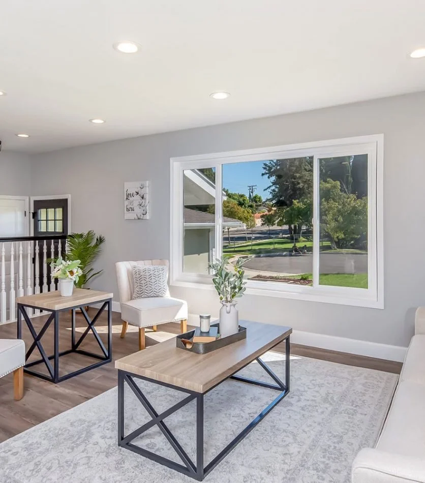 Bright living room with gray walls, wooden flooring, large window showing a street and trees outside, light-colored furniture, and modern decor.