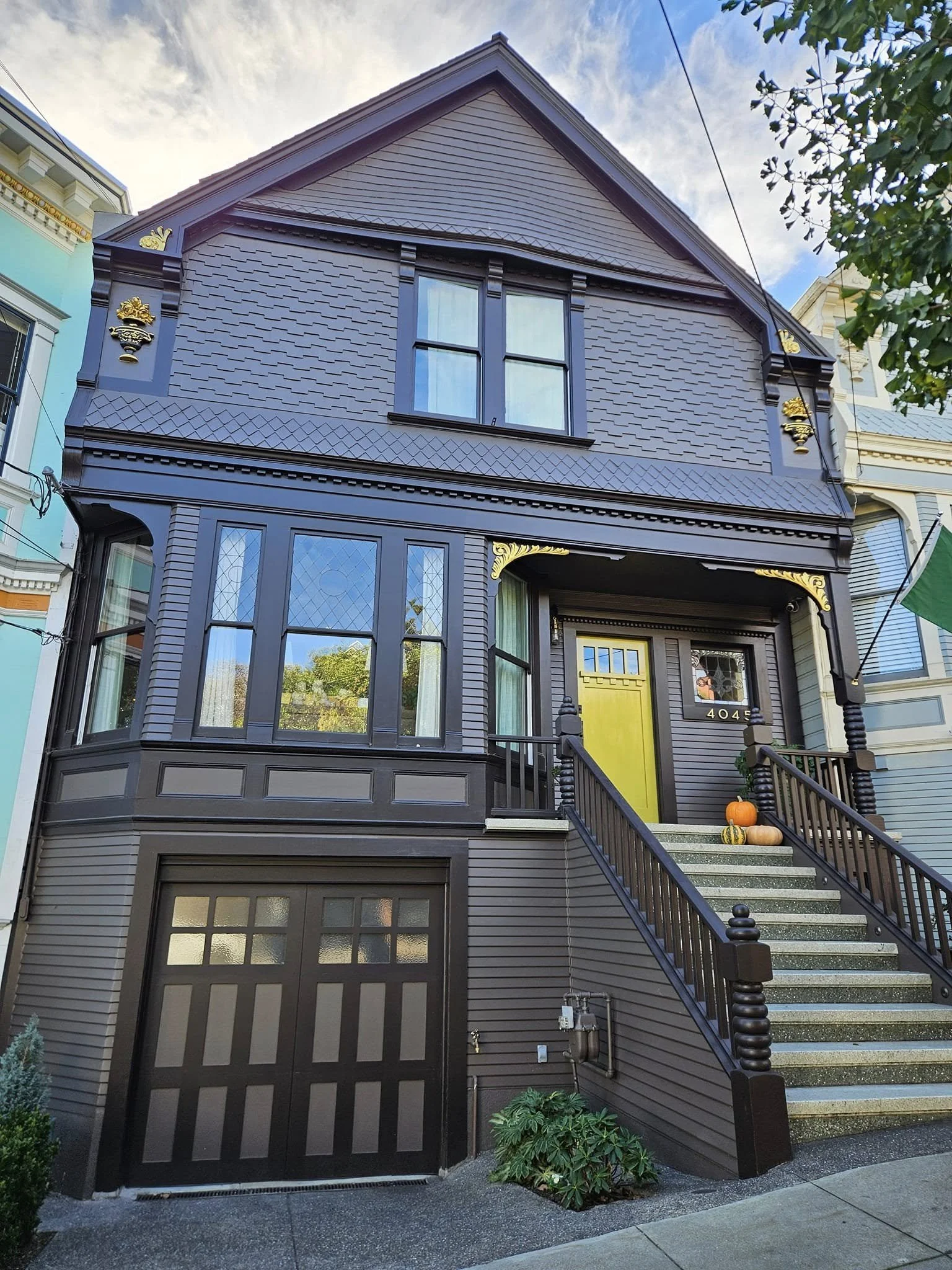 A dark-colored house with a decorative facade, featuring a large window on the upper level, a small window and a yellow front door on the lower level, with small pumpkins and gourds on the steps leading to the entrance, and trees and neighboring houses in the background.