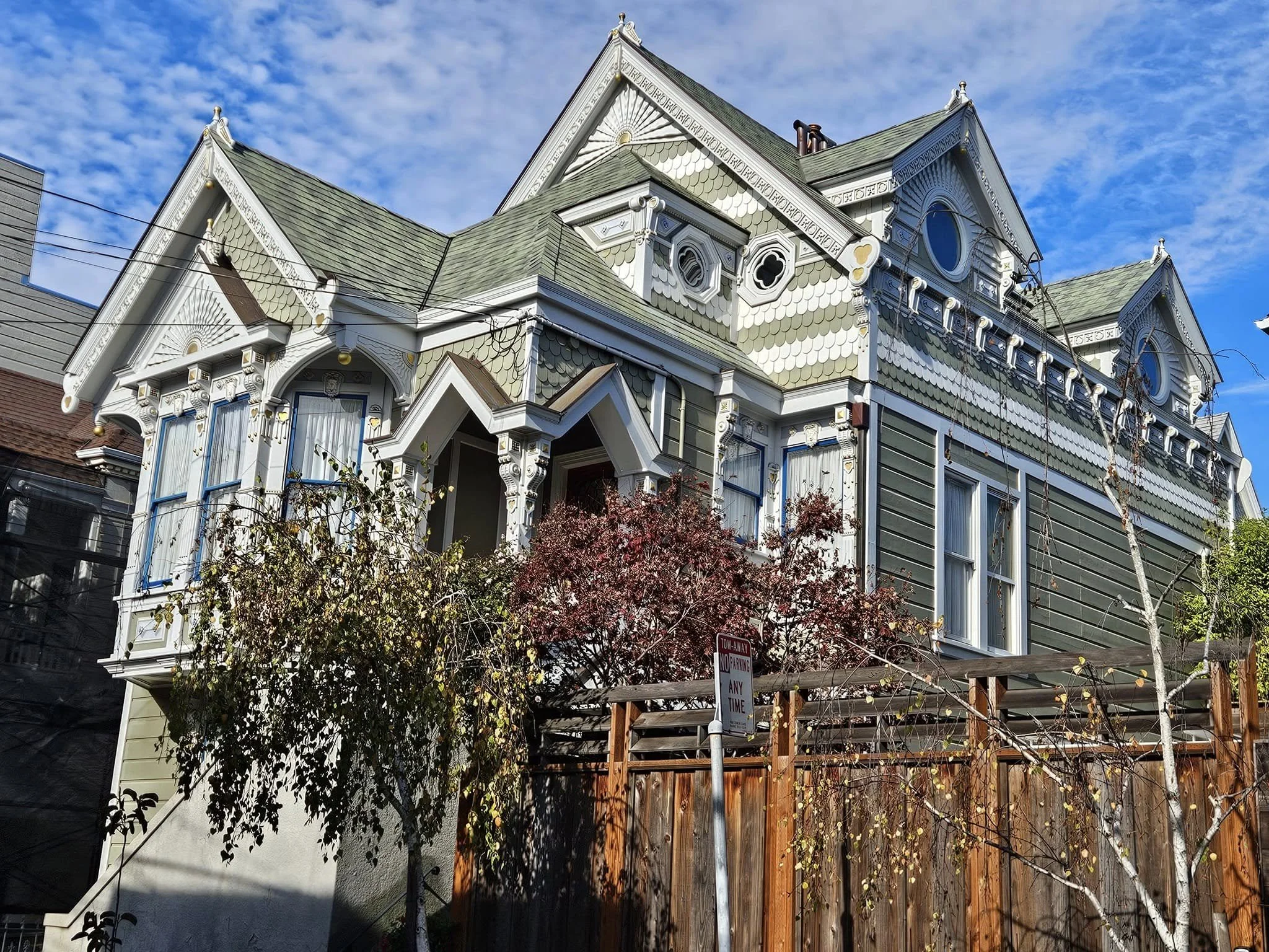 An ornate Victorian-style house with greenish-gray shingles and white decorative trim. The house has large bay windows, intricate woodwork, and a gabled roof against a partly cloudy blue sky. A construction scaffold is visible in front, and a red tree partially obscures the bottom of the house.