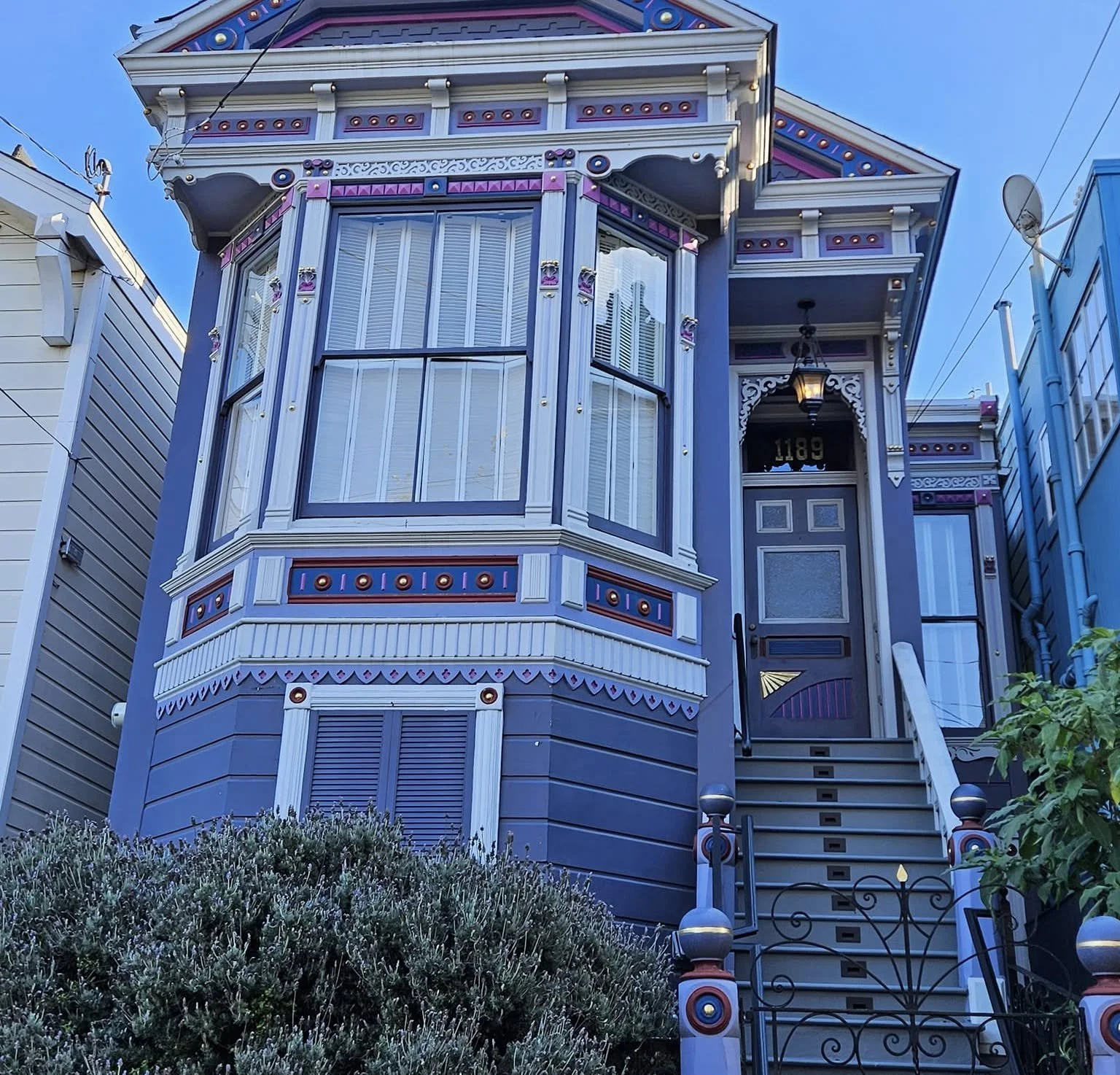 Colorful Victorian-style house painted purple, with ornate trim and large bay windows, situated on a small hill with stairway and plants in front.