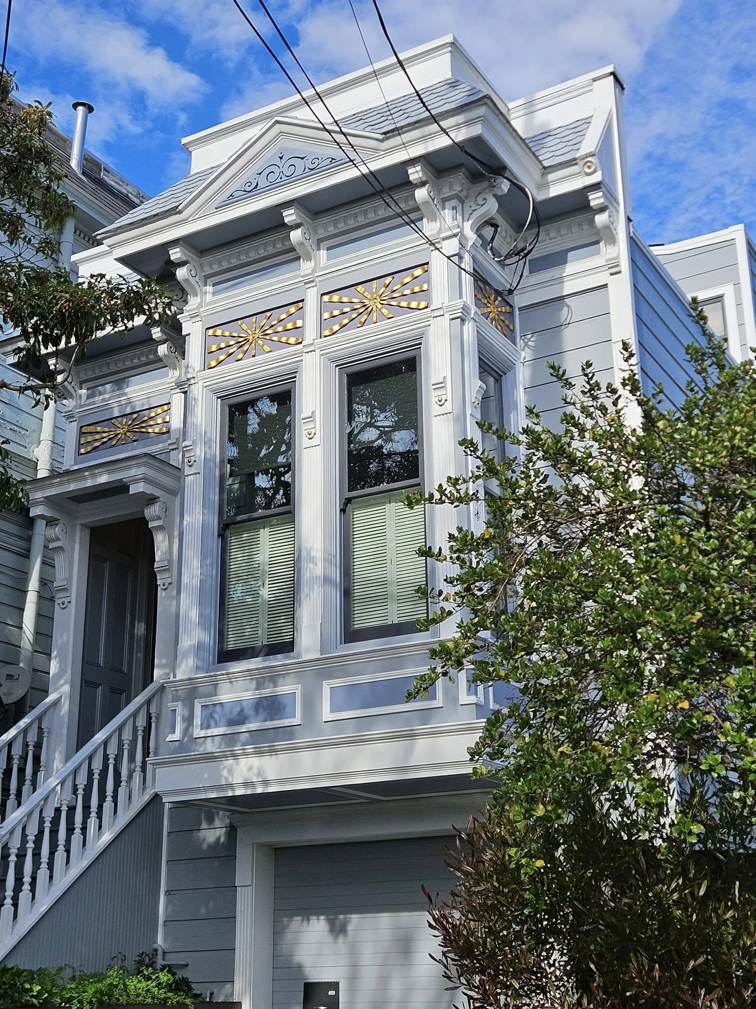 A white Victorian-style house with decorative gold accents on a balcony, large windows, and exterior moldings, with trees and a blue sky in the background.