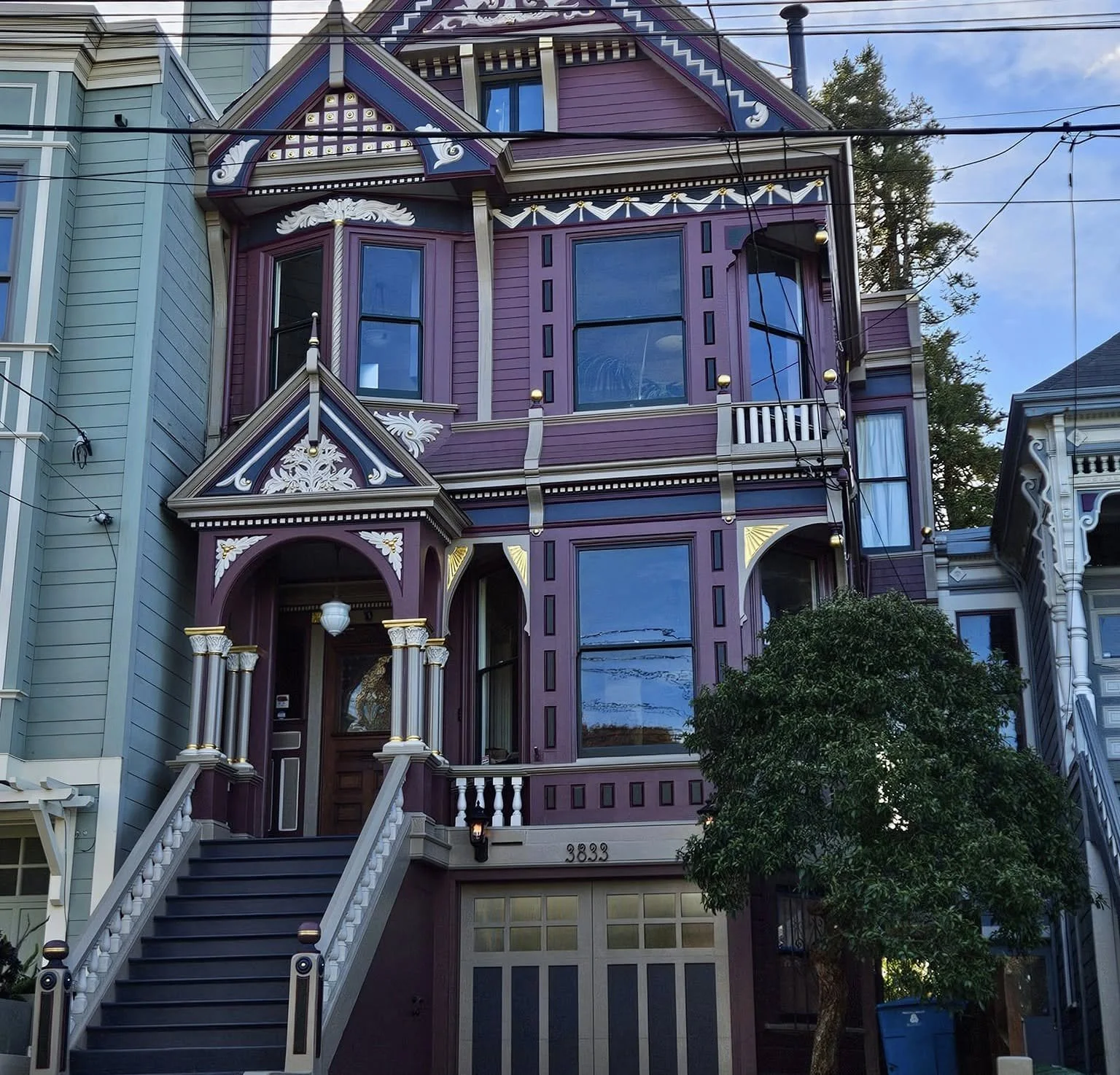 A colorful Victorian-style house painted purple with white and gold decorative trim, featuring large bay windows, a staircase leading to the front door, and a small porch. The house number 3833 is visible on the front.