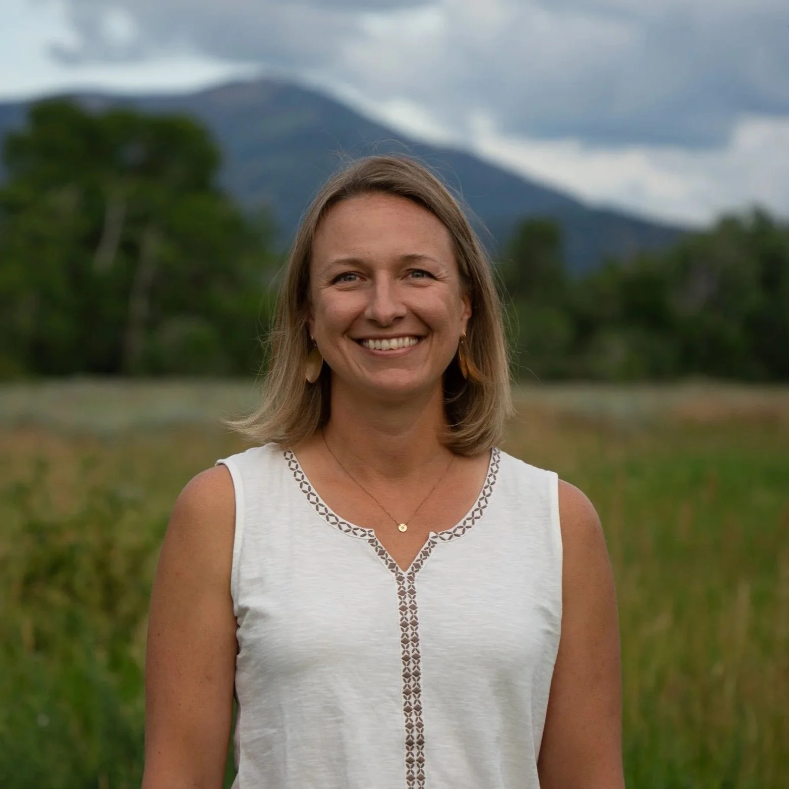 A woman with shoulder-length light brown hair smiling, standing outdoors in a field with green trees and mountains in the background, wearing a white sleeveless top with embroidered details.