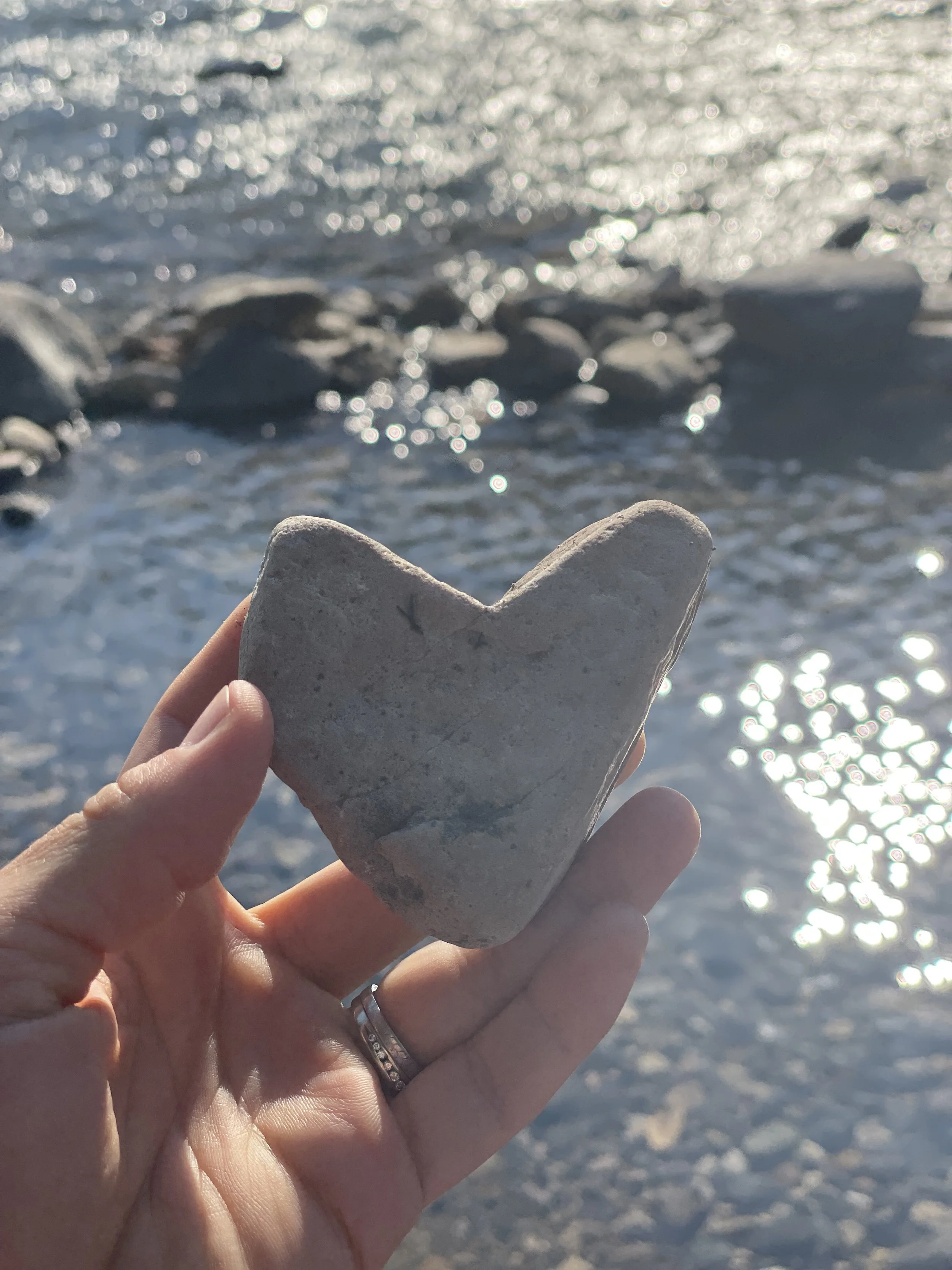 A person holding a heart-shaped rock in front of a sunlit body of water with rocks, creating a sparkling reflection.