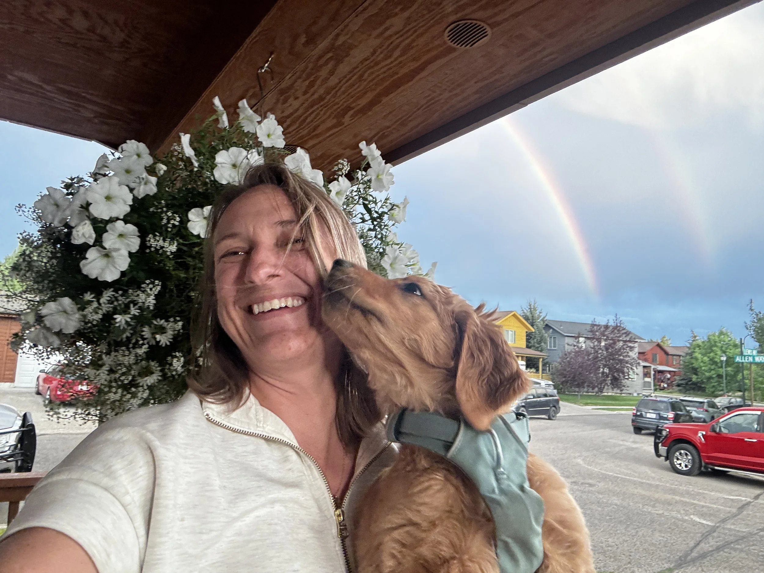 A woman with brown hair smiling and holding a golden retriever puppy outdoors on a porch with a flower arrangement behind her. In the sky, a rainbow is visible.